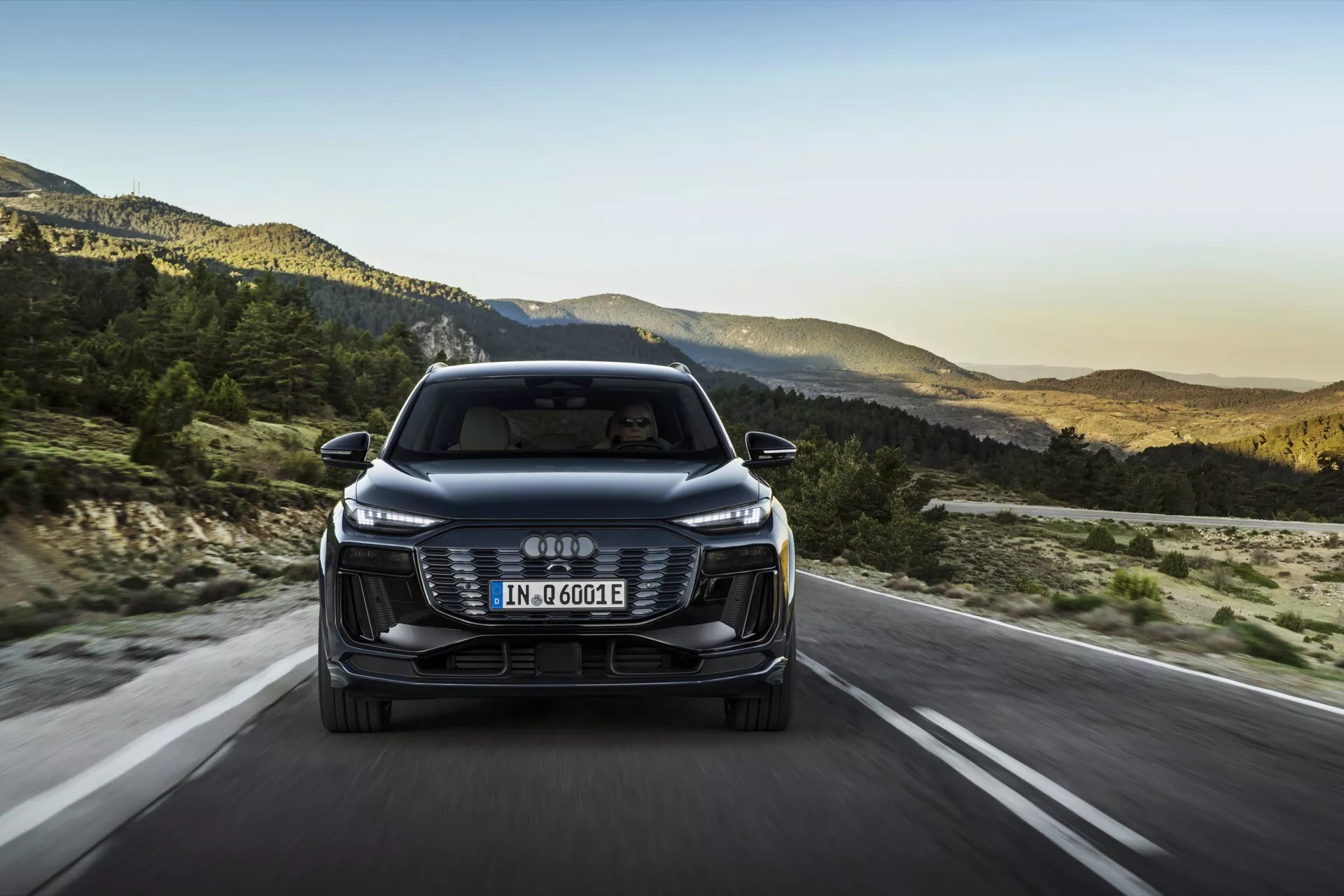 Vista frontal del Audi Q6 e-tron 2024, mostrando su parrilla delantera, faros y parachoques con el logo de los cuatro aros entrelazados. La matrícula de estilo europeo está montada en la parrilla, y la designación 'e-tron' indica su naturaleza eléctrica/híbrida. El coche está circulando por una carretera abierta con un paisaje montañoso de fondo, sugiriendo una ambientación de última hora de la tarde. Las características incluyen faros LED elegantes, parrilla estética, sensores/cámaras de asistencia al conductor, espejos aerodinámicos y un diseño eficiente.