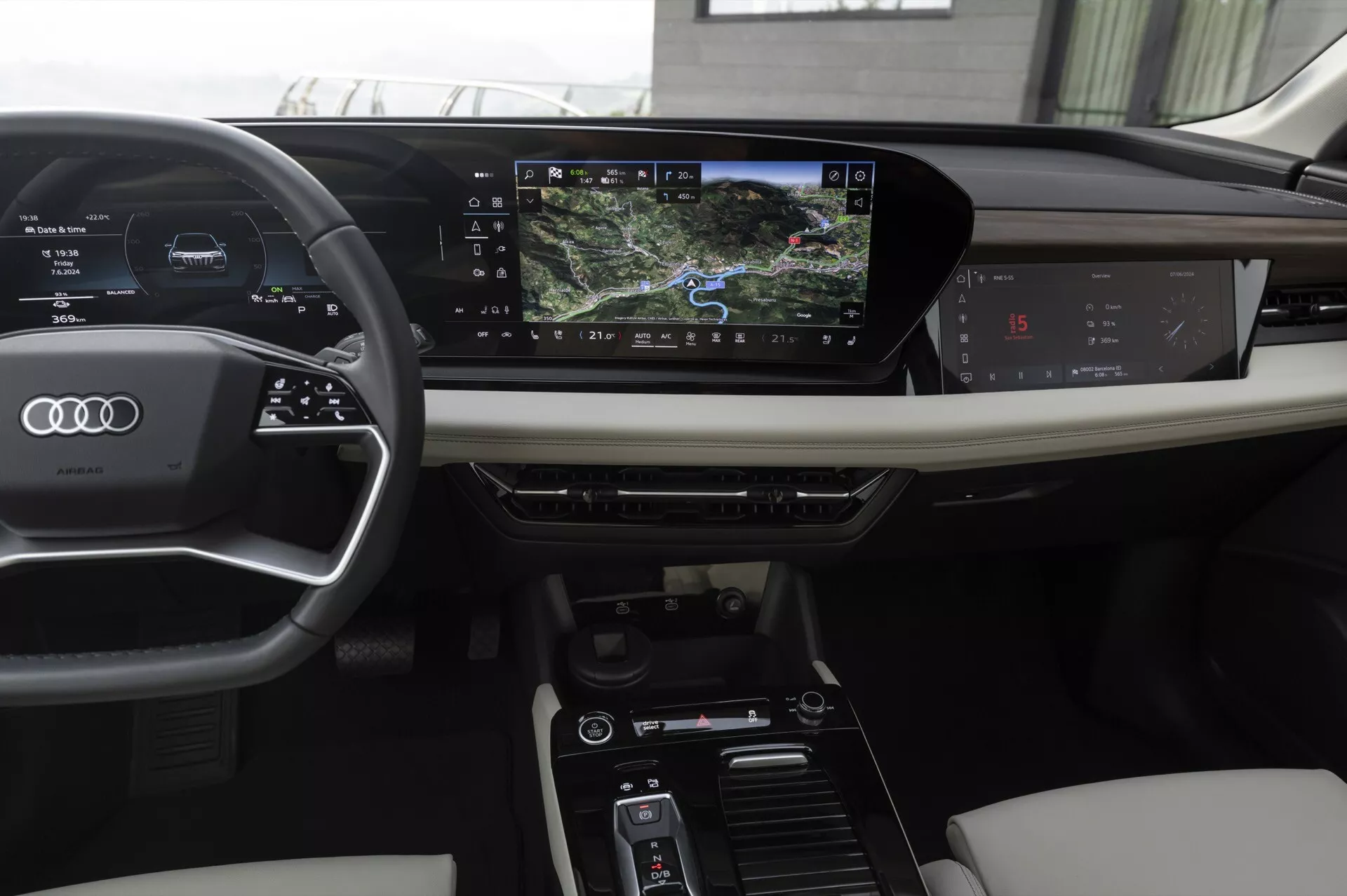 El interior de un moderno Audi Q6 e-tron 2024, visto desde la perspectiva del conductor. Las características incluyen un volante de tres radios envuelto en cuero con controles montados, un cuadro de instrumentos digital que muestra la velocidad y la navegación, y un sistema de infoentretenimiento con pantalla táctil horizontalmente orientada. La consola central incluye controles para configuración de clima y modos de conducción. Materiales lujosos como cuero y adornos de madera mejoran la estética.