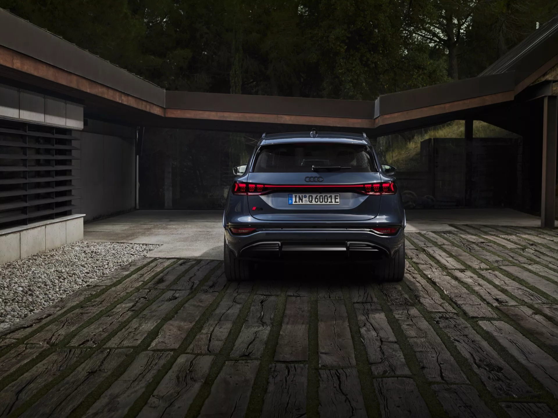 Vista de ángulo trasero del Audi Q6 e-tron 2024 que enfatiza su diseño limpio, elegante y moderno. Las características clave visibles son la franja de luz continua que conecta ambos faros traseros, la insignia del modelo y del tren motriz eléctrico, el parachoques trasero integrado con difusor deportivo, el parabrisas trasero inclinado hacia adelante que sugiere un SUV o crossover, las puntas de escape ocultas y la placa de matrícula centrada. El coche está aparcado en un camino de adoquines con un toque de un garaje contemporáneo en el fondo, indicando un estilo de vida lujoso y eco-consciente.