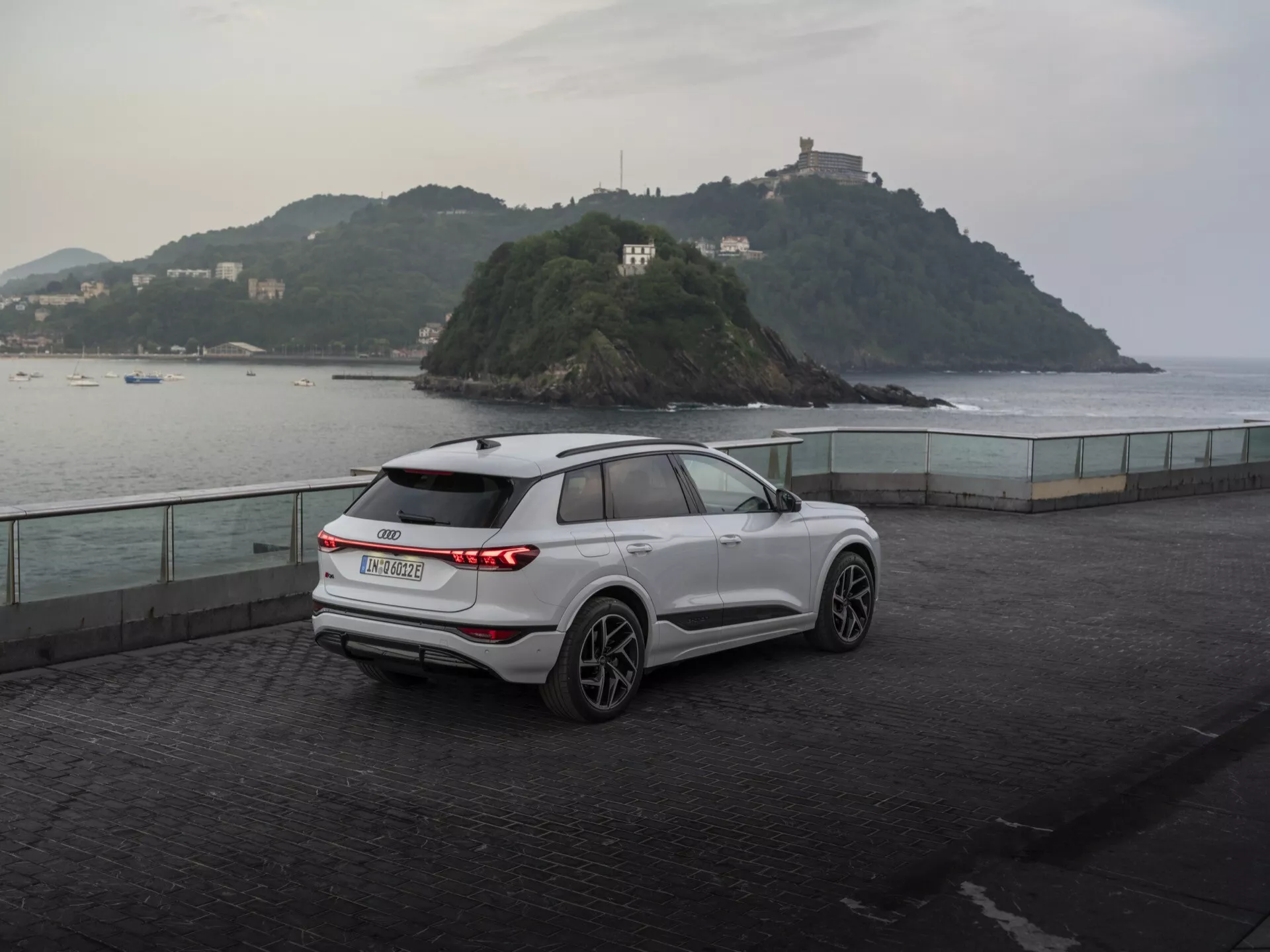 Vista trasera tres cuartos de un moderno SUV Audi Q6 e-tron 2024 blanco estacionado en un paseo marítimo al anochecer, mostrando sus luces traseras LED horizontales, emblema del fabricante, limpiaparabrisas trasero, llantas de aleación de cinco radios, espejos con indicadores de giro integrados, barras de techo y líneas esculpidas de la carrocería contra un fondo sereno con una pequeña isla y un edificio, destacando una mezcla de utilidad y rendimiento.
