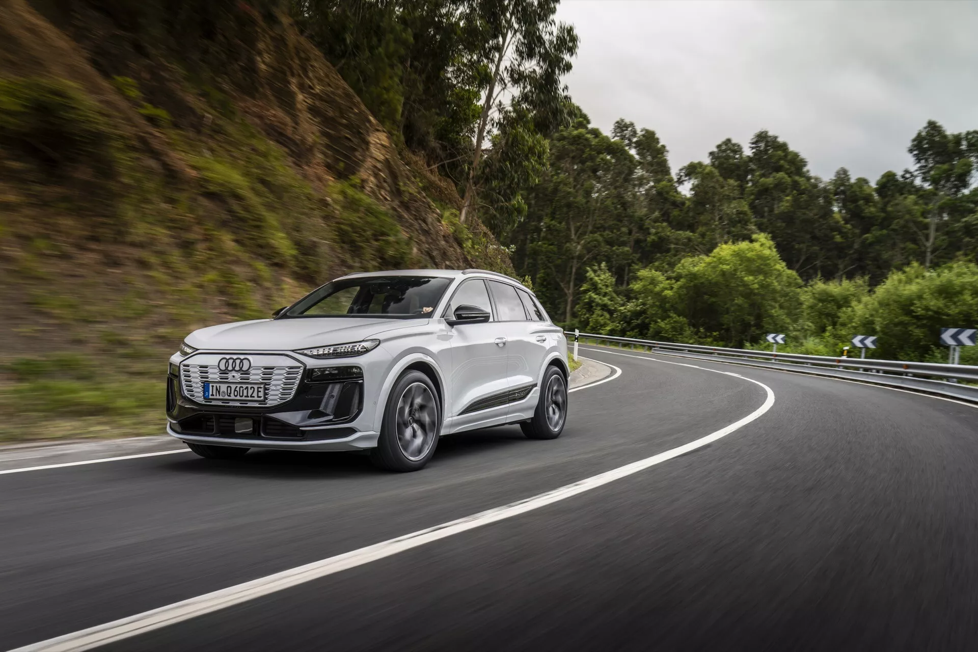 La imagen captura una vista lateral dinámica de un SUV blanco moderno, probablemente un modelo Audi Q6 e-tron 2024, viajando por una carretera bordeada por una zona boscosa. El enfoque está en la parrilla frontal, los faros, el perfil lateral, las ruedas y la aerodinámica de los espejos laterales del vehículo. Los faros son elegantes con tecnología LED, lo que sugiere una iluminación avanzada. Las llantas de aleación con un diseño de múltiples radios sugieren capacidades de alto rendimiento. El movimiento del automóvil en una carretera curva resalta sus capacidades de manejo y posibles sistemas avanzados de asistencia a la conducción. El fondo desenfocado enfatiza la velocidad y la eficiencia en la navegación de escenarios desafiantes.