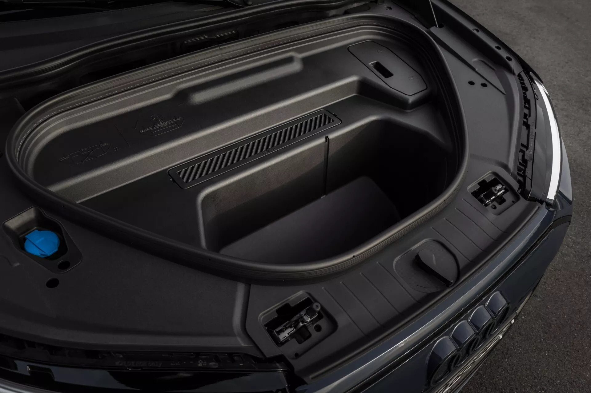 Toma en primer plano desde un ángulo alto del compartimento trasero del Audi Q6-e-Tron 2024, que muestra las características interiores, incluidos los paneles de plástico moldeado para durabilidad, enchufes de accesorios de potencia en el lado derecho, almacenamiento empotrado en la parte inferior y un tapón azul a la izquierda, indicativo de tecnología automotriz avanzada y un acabado premium y elegante.