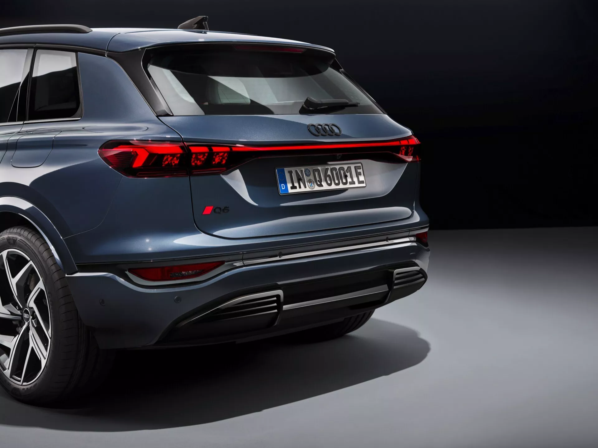 Vista de tres cuartos trasera del Audi Q6 e-tron 2024 que muestra luces traseras LED, parachoques trasero deportivo con salidas de escape integradas, rueda aleación multiradio, acabado brillante metálico oscuro, matrícula trasera europea insinuando electrificación, y moldura cromada en ventanas en un entorno de iluminación controlada.