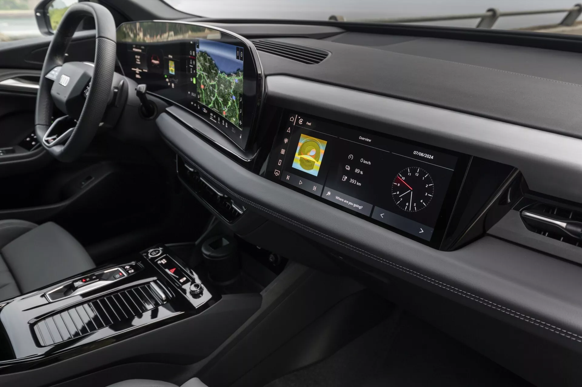 Vista del interior del Audi Q6 e-tron 2024, enfocándose en el tablero y consola central desde un ángulo ligeramente elevado. A la izquierda se muestra el volante, indicando configuración para conducir en el lado izquierdo, mientras que la consola central presenta una palanca de cambios, varios controles y una gran pantalla táctil. El clúster de instrumentos digital muestra métricas de conducción. El tablero elegante se extiende horizontalmente con ventilaciones y otro elemento de diseño a la derecha. La tecnología avanzada es evidente con la pantalla táctil central mostrando información de navegación y multimedia, una pantalla proyectada visible sobre el volante y botones táctiles para el control del clima. El diseño enfatiza el minimalismo y materiales premium, con costuras de cuero y acentos metálicos que realzan la sensación de lujo.