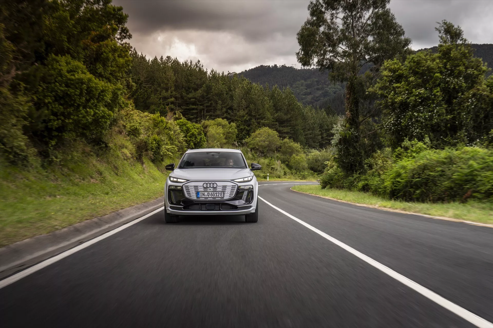 Audi Q6-e-tron 2024 captado en movimiento desde un ángulo frontal en una carretera de dos carriles bordeada de vegetación. La foto muestra la parrilla, los faros, el capó y el parachoques delantero del vehículo, resaltando su rendimiento y diseño dinámicos. El coche cuenta con faros LED distintivos, un emblema de lujo prominente en la parrilla y líneas aerodinámicas, todo bajo un clima nublado con condiciones de carretera seca.