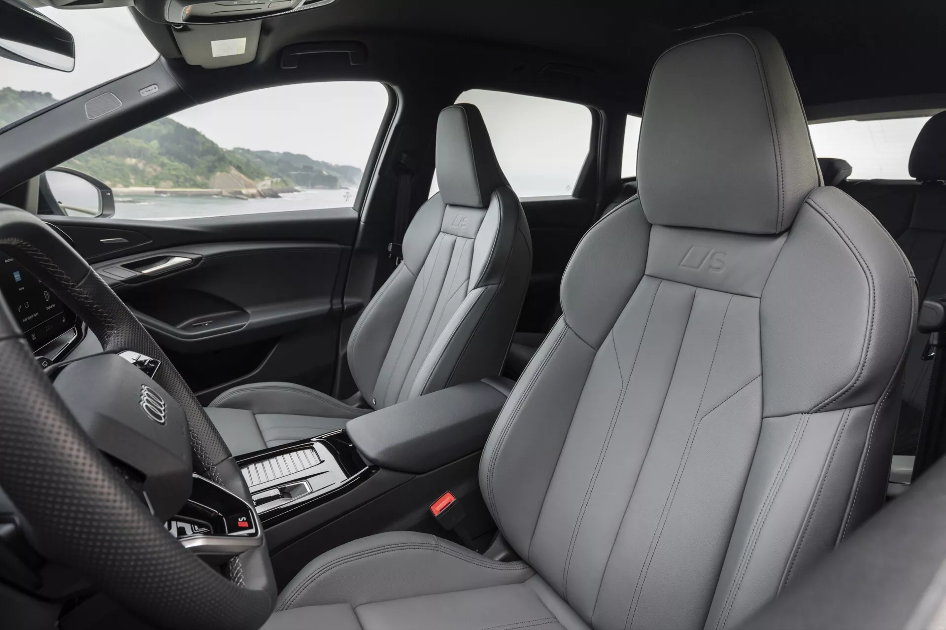 Vista interior del Audi Q6 e-Tron 2024 que muestra los asientos delanteros de cuero lujoso con un patrón de costura prominente, logotipo en relieve y diseño contemporáneo. La fotografía destaca el tablero con un volante con controles integrados y un clúster de instrumentos digital que no está iluminado. La consola central, que alberga la palanca de cambios y botones ordenadamente dispuestos, es parcialmente visible. La imagen captura los reposabrazos tapizados de las puertas con controles para espejos, ventanas y posiblemente configuraciones de asiento. La iluminación natural resalta la comodidad y la alta calidad de los materiales en la cabina.