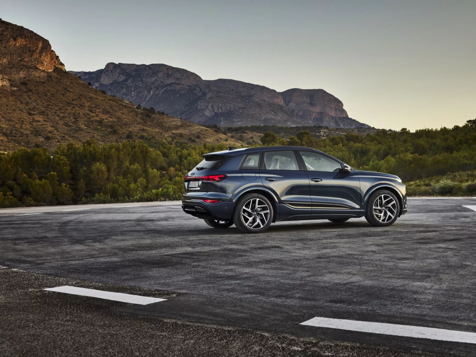 Imagen de un SUV Audi Q6-e-tron 2024 estacionado en un aparcamiento al aire libre con un paisaje natural, posiblemente montañoso, de fondo. La foto captura una vista trasera de tres cuartos, destacando tanto los elementos de diseño laterales como traseros. El lado del conductor es claramente visible, mostrando las líneas de diseño, las ventanas traseras y el diseño de las ruedas. La línea del techo inclinada sugiere un diseño deportivo tipo coupé. Las luces traseras LED modernas, los sensores o reflectores visibles en el parachoques trasero y el esquema de color dual enfatizan la altura y la postura. Las grandes llantas de aleación y los neumáticos sustanciales indican un nivel de acabado superior con una estética deportiva. El vehículo parece estar cerca de la cámara, proporcionando una vista detallada. Las características notables incluyen iluminación LED, sensores de estacionamiento, ruedas y neumáticos de alto rendimiento, un diseño aerodinámico, y posiblemente sistemas avanzados de asistencia al conductor y tracción en las cuatro ruedas.