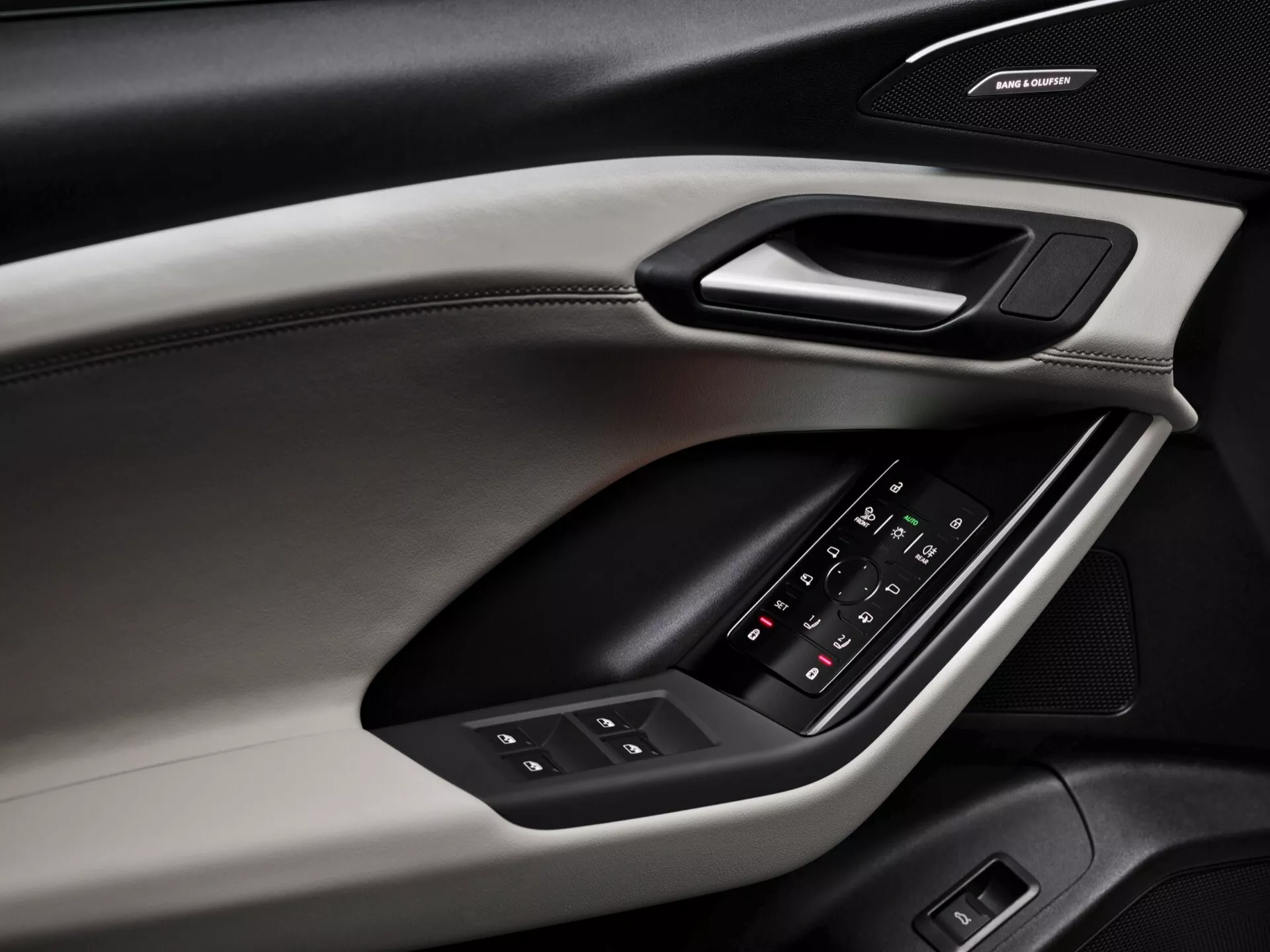 Vista de cerca del panel de la puerta interior del Audi Q6 e-tron 2024, mostrando elementos de diseño y características. El panel de la puerta, capturado desde un ángulo diagonal, revela diferentes materiales y texturas, incluyendo una sección superior suave al tacto con costuras visibles, un borde duro que alberga una manija ergonómica y un reposabrazos acolchado con panel de control retroiluminado para ventanas y ajustes de espejos laterales. Debajo del reposabrazos, un sistema de altavoces con marca indica un sistema de audio premium. El esquema de color incluye un diseño de dos tonos, mejorando las cualidades estéticas y funcionales.