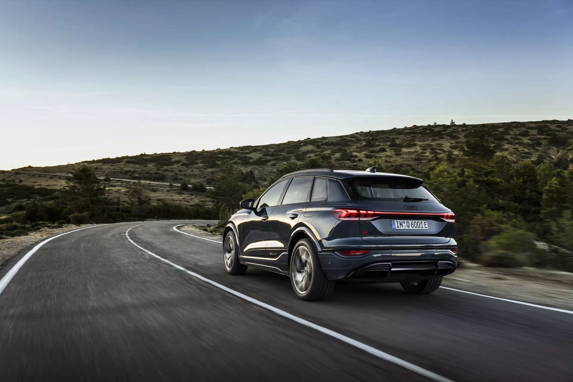 La imagen captura un Audi Q6 e-tron 2024 desde un ángulo trasero de tres cuartos, destacando el lado derecho y la parte trasera del vehículo. El coche, que parece estar en movimiento en una carretera sinuosa, muestra sus elementos de diseño, incluidas las luces traseras, el parachoques trasero, las salidas de escape, las llantas de aleación y parte del perfil lateral con la puerta trasera y la ventana del pasajero. El fondo borroso implica velocidad, y la iluminación natural sugiere que la foto fue tomada durante el día, en un entorno rural o semi-rural con vegetación escasa. Se sugieren características avanzadas como luces traseras LED, un alerón trasero o escapes dobles, aunque los detalles tecnológicos más finos son indistinguibles debido al desenfoque de movimiento.
