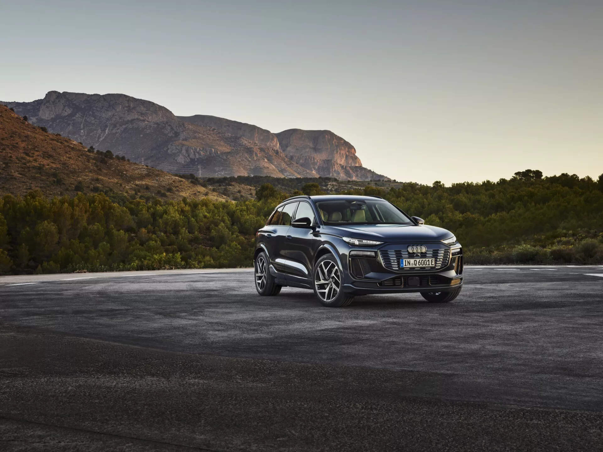 SUV de lujo Audi Q6 e-tron 2024 en ángulo frontal de tres cuartos mostrando una parrilla delineada en cromo, faros LED modernos, grandes ruedas de aleación de múltiples radios, espejos laterales con señales de giro integradas, ventanas tintadas y acabado de pintura brillante, estacionado sobre asfalto con montañas al fondo al amanecer o al atardecer.