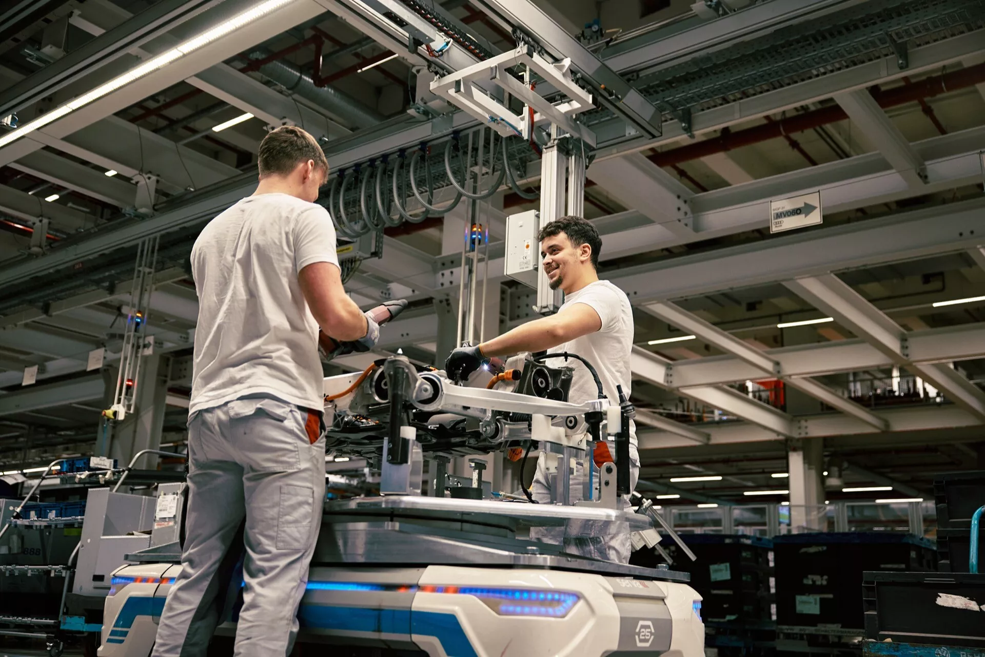 Entorno industrial con dos trabajadores ensamblando o manteniendo equipos mecánicos en el suelo de una fábrica, bajo iluminación artificial, con un punto de vista elevado que enfatiza el ambiente de manufactura