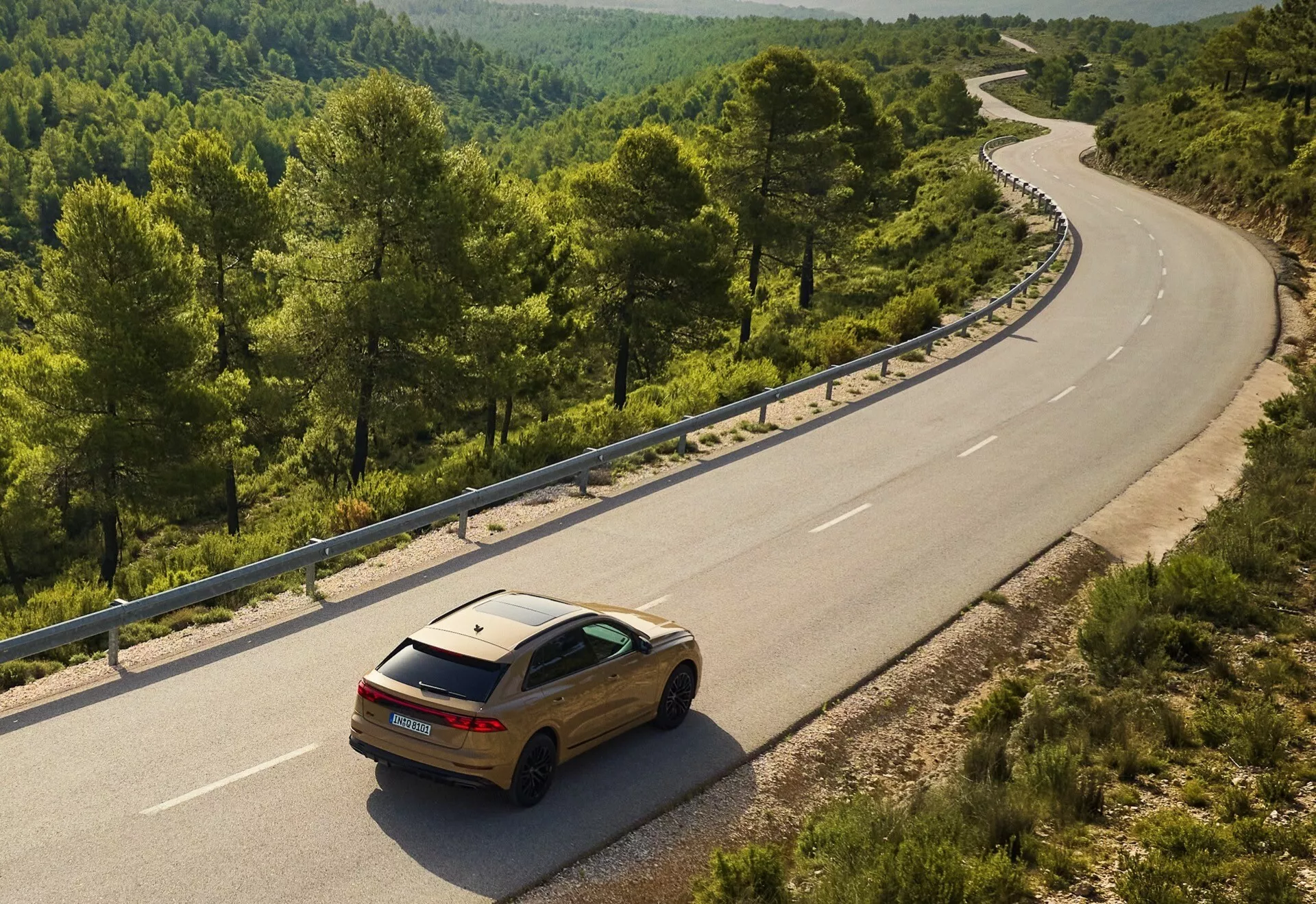 Audi Q8 2023 (4M-Facelift) de color oro en una carretera sinuosa de dos carriles en un entorno rural o forestal, visto desde un ángulo elevado mostrando su diseño trasero y lateral contra el paisaje natural