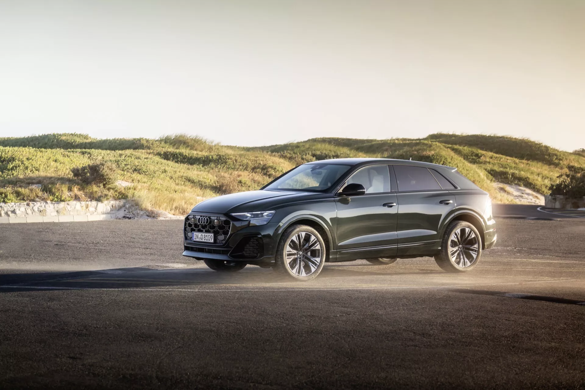 Crossover moderno Audi Q8 2023 (restyling 4M) en exteriores durante la hora dorada, mostrando faros LED avanzados, parrilla de lujo con características de seguridad, llantas de aleación grandes orientadas al rendimiento, pintura metálica y líneas de diseño deportivas contra un fondo de asfalto y colinas suaves, destacando capacidades versátiles y diseño