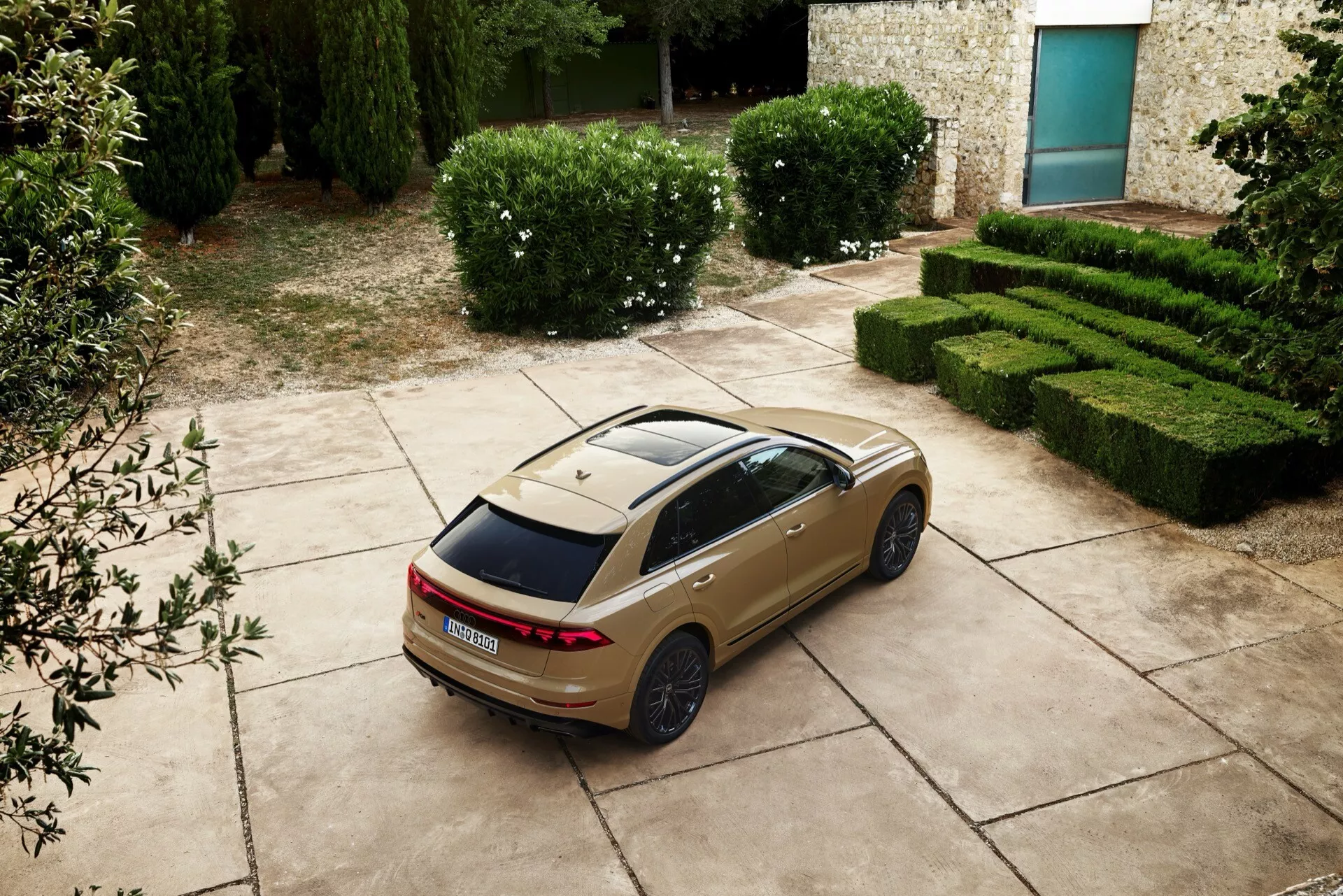 Sedán Audi Q8 2023 (4M-facelift) en vista trasera y lateral desde un ángulo alto mostrando luces traseras LED, techo solar panorámico y llantas deportivas, aparcado con un fondo escénico de setos y un edificio de piedra