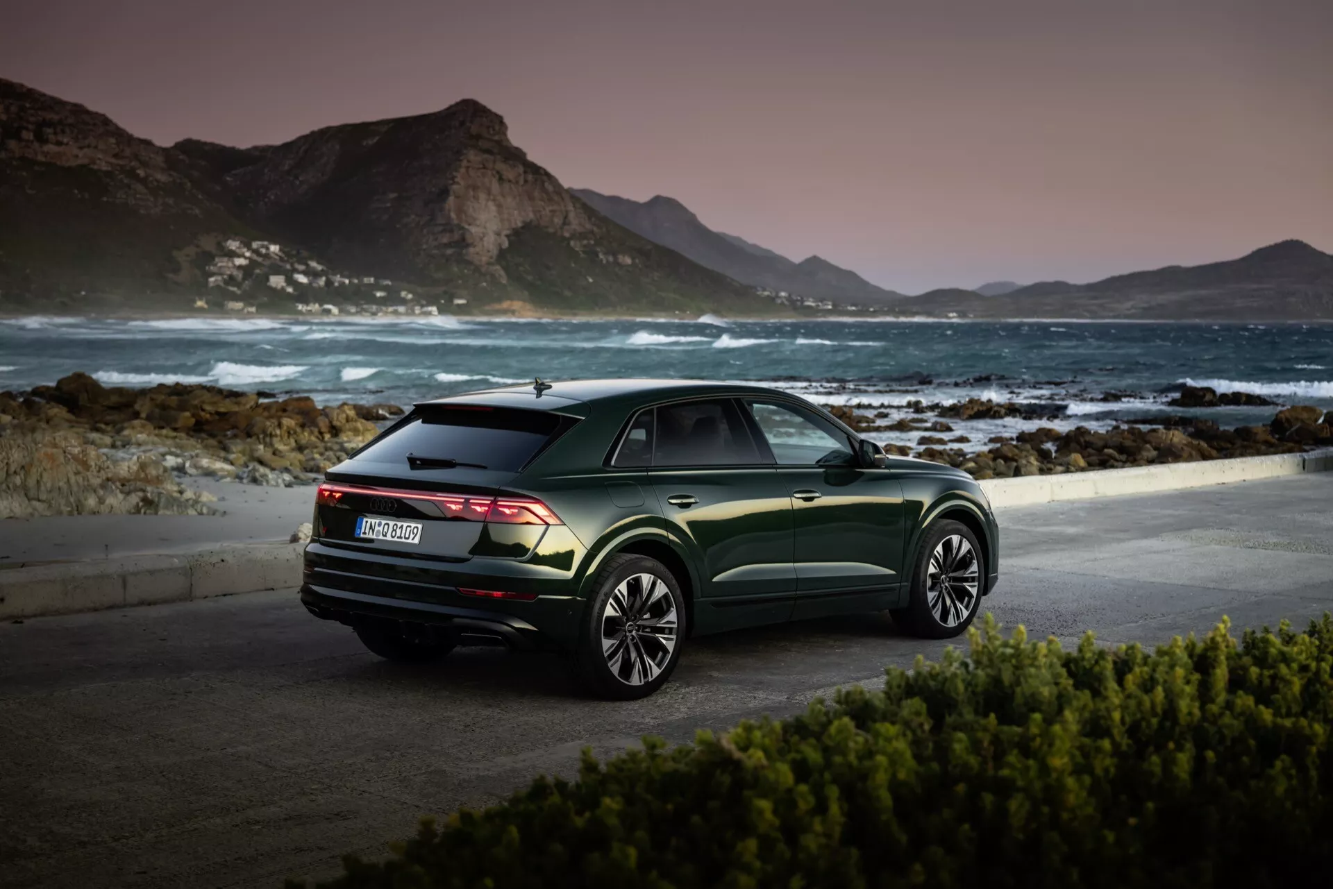 Audi Q8 2023 (4M-Facelift) estacionado en un pintoresco lugar junto al mar al amanecer o al atardecer, mostrando su vista de tres cuartos traseros, acabado en verde metálico oscuro, y ruedas de aleación de múltiples radios frente a un sereno fondo montañoso