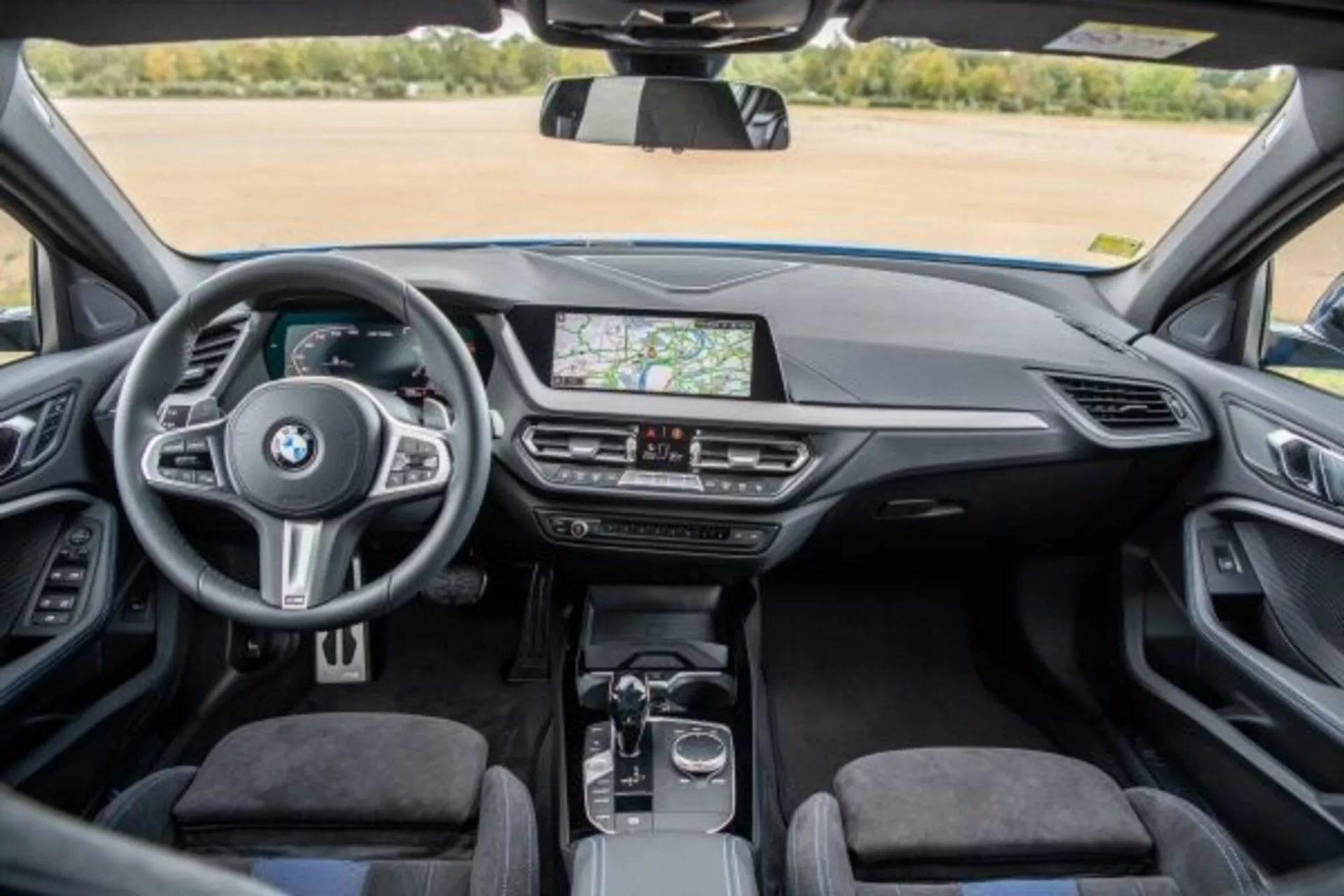 Vista interior del BMW Serie 1 M Performance 2019 desde el asiento trasero mostrando el tablero, la consola central y el volante con controles montados, sistema de información y entretenimiento con pantalla táctil mostrando navegación GPS, controles de clima, palanca de cambios automática, dial rotatorio y panel táctil en la consola central, tapicería premium y cabina clara y bien iluminada