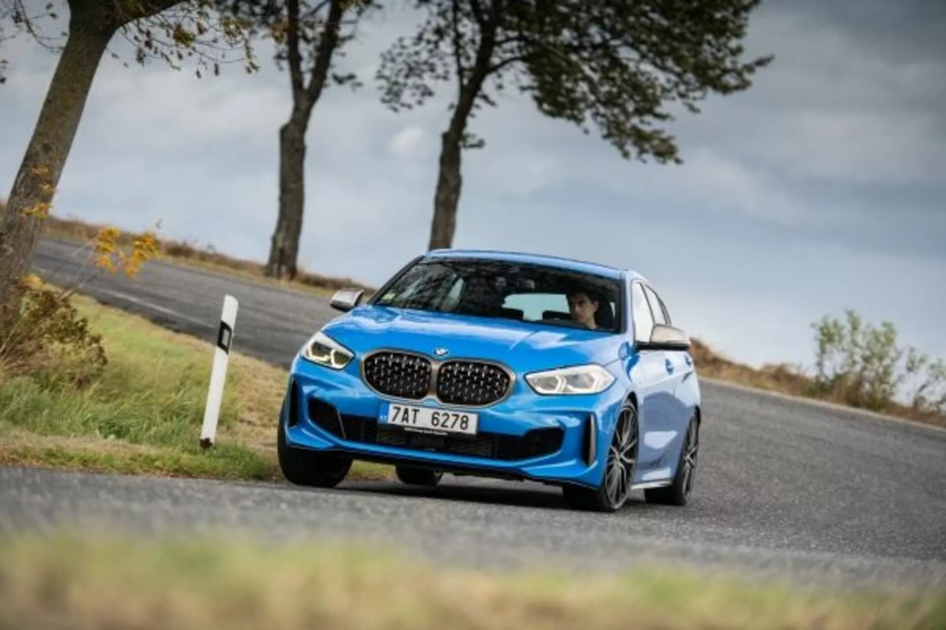 Fotografía dinámica desde un ángulo bajo de un moderno sedán deportivo azul BMW Serie 1 M Performance 2019 en movimiento en una carretera rural, mostrando su diseño, la distintiva parrilla en forma de riñón, faros LED agresivos, parachoques delantero esculpido con entradas de aire y ruedas de aleación