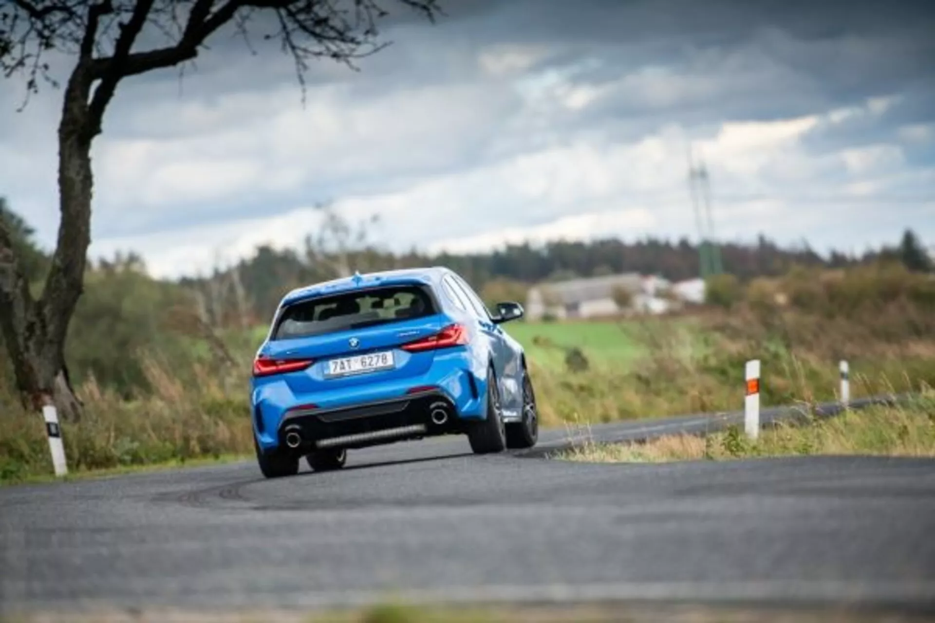 BMW Serie 1 M Performance de 2019 en azul conduciendo por un camino rural, vista trasera de tres cuartos destacando diseño aerodinámico con alerón trasero, luces traseras LED modernas y sistema de doble escape