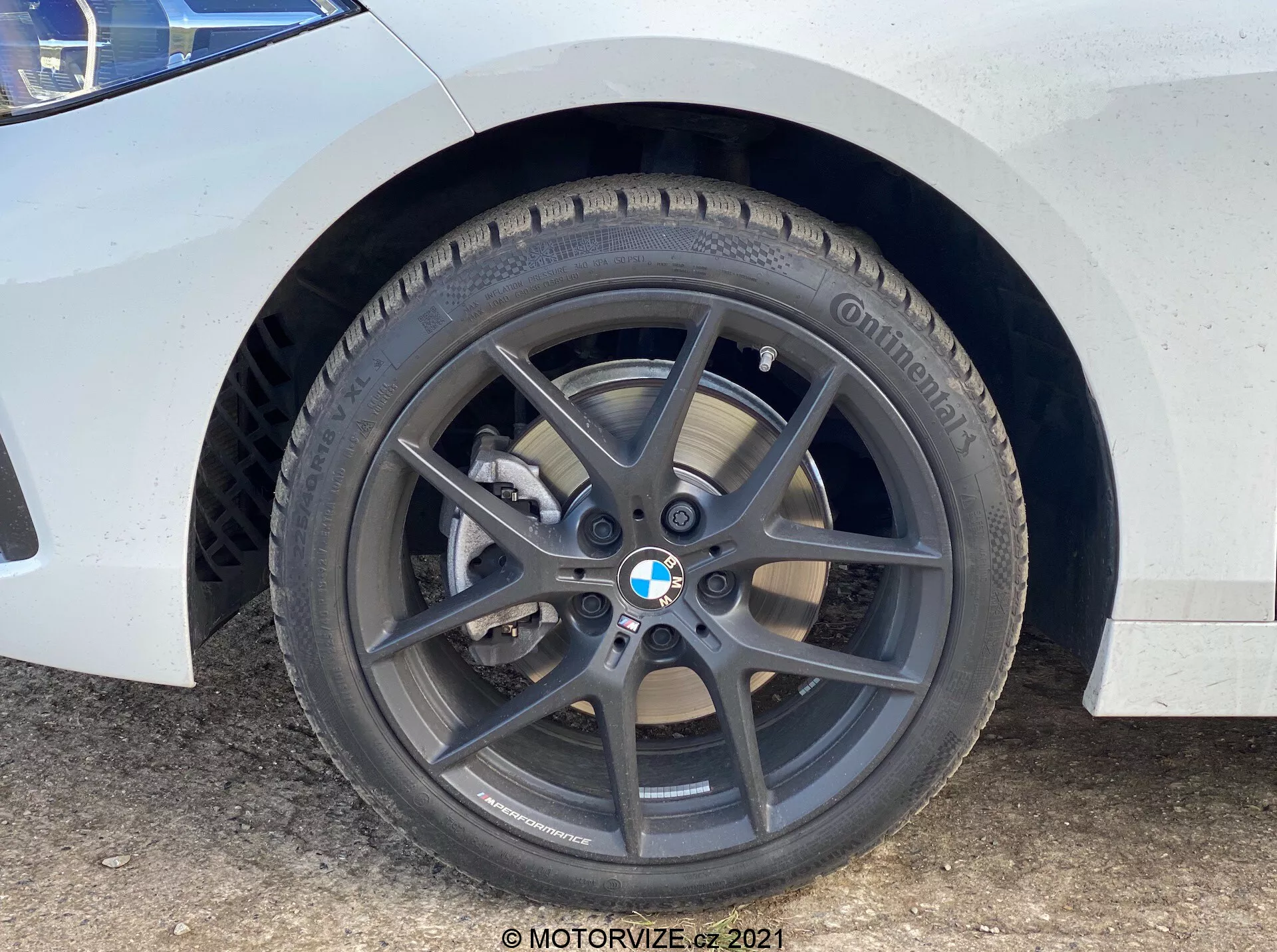 Primer plano de la rueda delantera y guardabarros del BMW 1er-Reihe 2019 (BMW-116i). La perspectiva a nivel del suelo destaca la rueda de aleación de cinco radios en gris oscuro con el emblema de BMW, neumático Continental con banda de rodadura visible, disco de freno ventilado y pinza de freno de un solo pistón en color plateado. La pintura blanca con manchas de suciedad y agua indica un uso regular.
