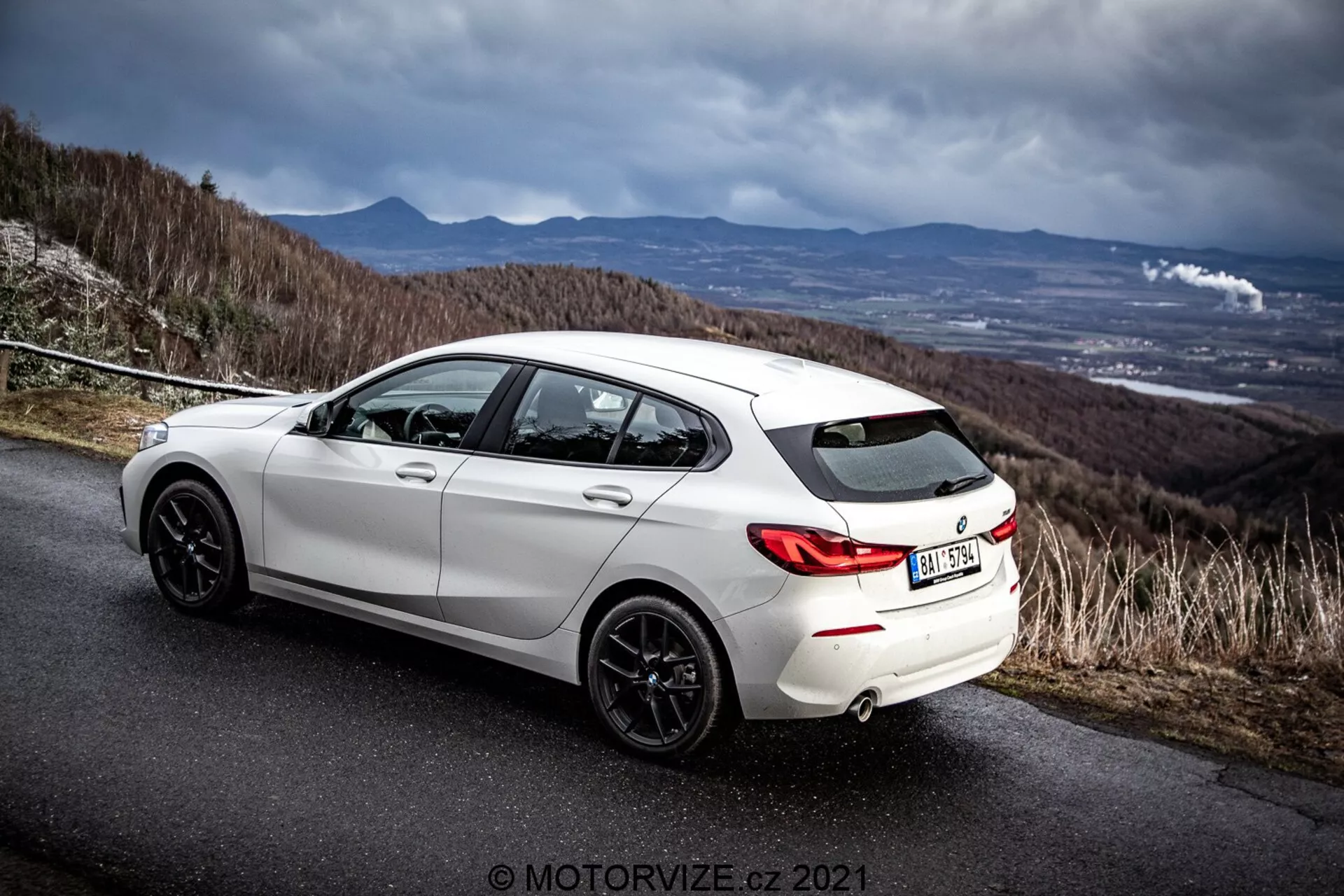 Vista trasera lateral en ángulo de un hatchback blanco BMW 116i 2019 en una carretera de asfalto con un cielo dramático y nublado. La imagen destaca el diseño trasero del vehículo, los arcos de las ruedas traseras, las llantas de aleación de color oscuro, el sistema de escape dual y el perfil lateral, incluidos los puertas traseras y delanteras del pasajero. El entorno sugiere una carretera de montaña o colina, enfatizando los atributos deportivos y dinámicos del coche. Marca de agua del fotógrafo 'MOTORWIZE.cz 2021' presente.