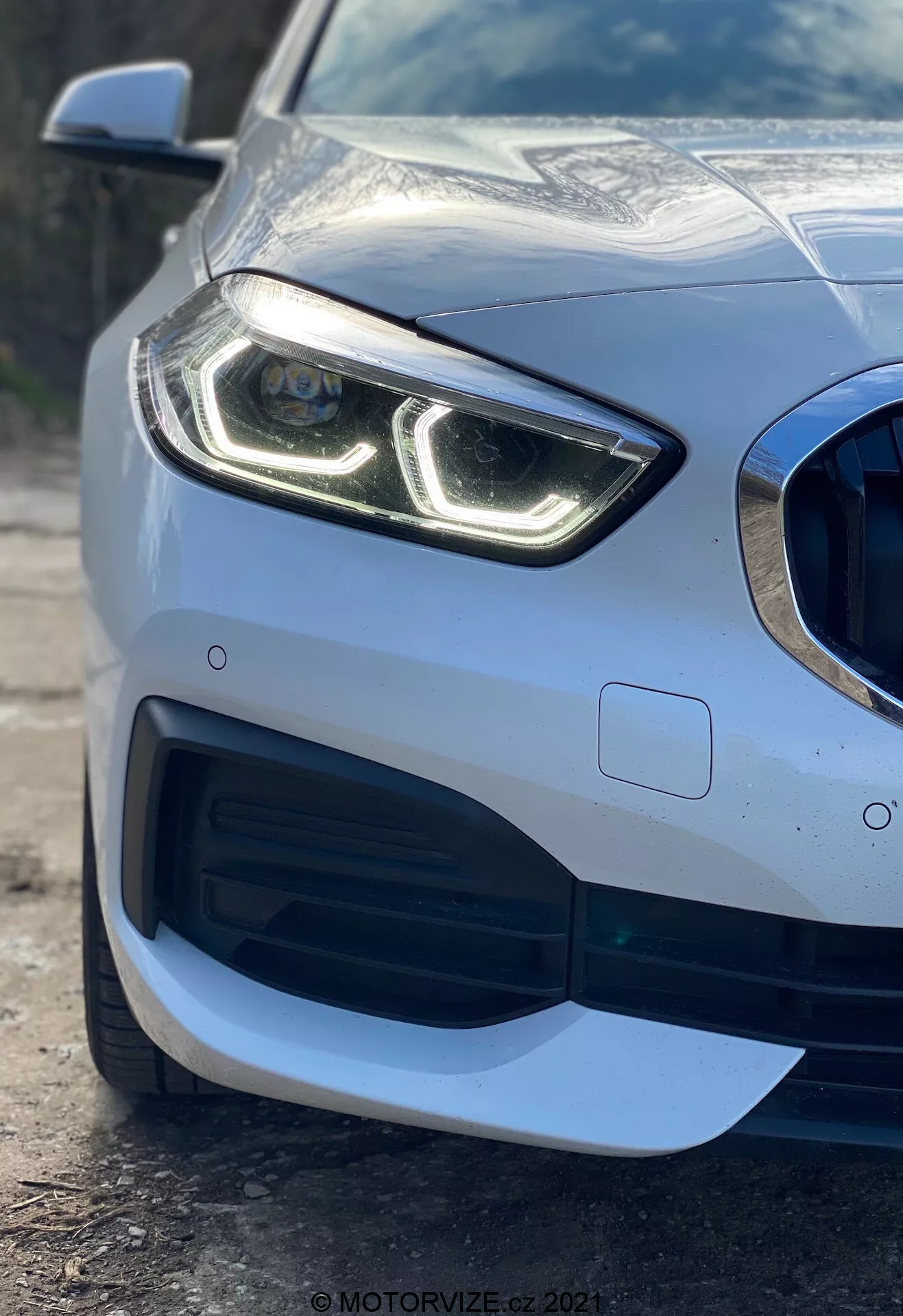 Vista de cerca de la sección frontal izquierda de un coche BMW Serie 1 2019 (BMW 116i) destacando su faro moderno con luces diurnas LED distintivas, parrilla con patrón de panal y gran toma de aire, diseño aerodinámico sofisticado en el parachoques delantero y sensor de aparcamiento incrustado en el parachoques, mostrando el acabado de pintura metálica premium y la carrocería reflectante.