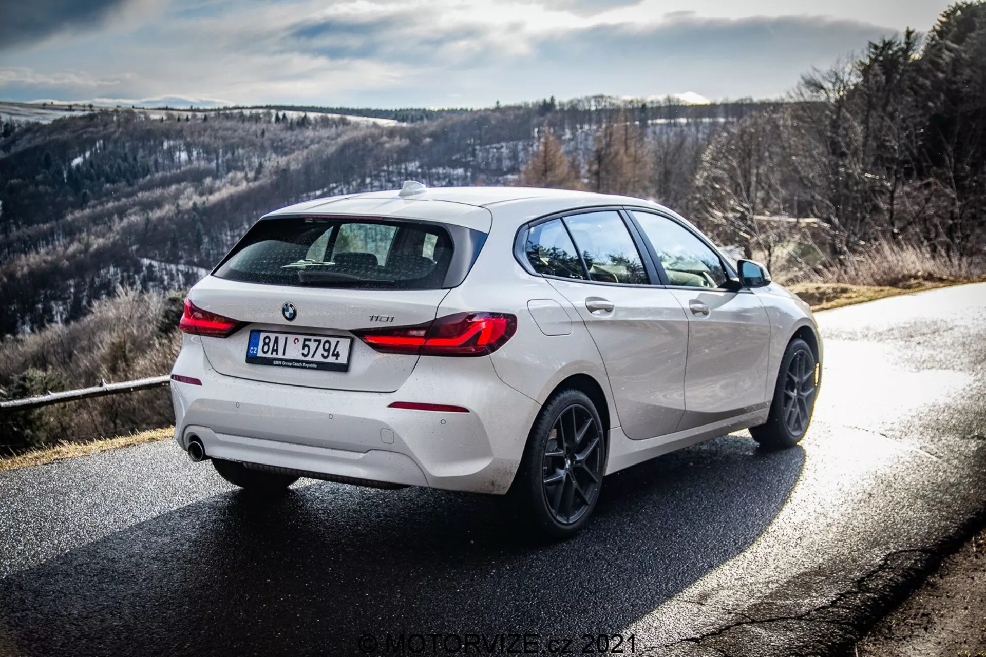Vista trasera de tres cuartos dinámica de un BMW 116i 2019 blanco, mostrando su lado derecho y trasero bajo la luz del día con claros rayos de sol y sombras. La imagen resalta los detalles de diseño, incluyendo acentos negros en el difusor trasero, llantas y carcasas de espejos laterales, complementados por ruedas de aleación y un sistema de escape doble, sugiriendo una variante más deportiva. La carretera se curva a la derecha con un fondo montañoso o colinoso, ideal para los entusiastas de la conducción.