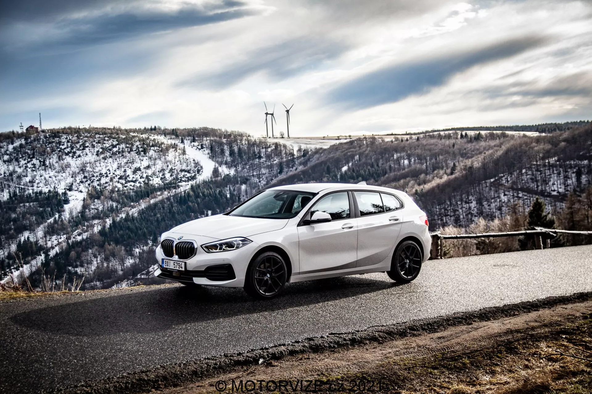 Un coche hatchback BMW blanco, probablemente un BMW 1er-Reihe 2019 (BMW 116i), está aparcado en una carretera asfaltada curva rodeada de un paisaje montañoso y nevado con turbinas eólicas. La foto, tomada desde un ángulo frontal lateral, resalta la parrilla delantera, los faros, las líneas aerodinámicas y las llantas de aleación del coche bajo un cielo nublado.