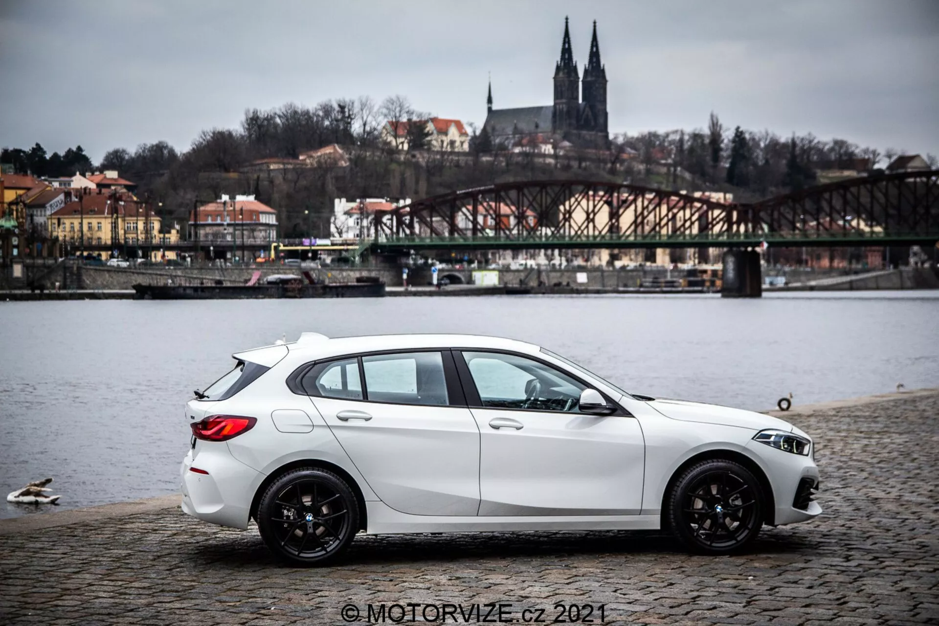 Un hatchback BMW Serie 1 de 2019 (BMW-116i) en blanco, estacionado sobre una superficie de adoquines cerca de un cuerpo de agua con patos, mostrando un perfil lateral con vista trasera parcial. El coche tiene líneas de carácter esculpidas dinámicamente, una línea de techo descendente, ruedas de aleación negras de múltiples radios y faros y espejos laterales visibles. El fondo incluye un puente, edificios en una colina y elementos arquitectónicos que sugieren un entorno europeo. La iluminación es difusa bajo un cielo nublado y la imagen incluye una marca de agua © MOTORVIZE.cz 2021.