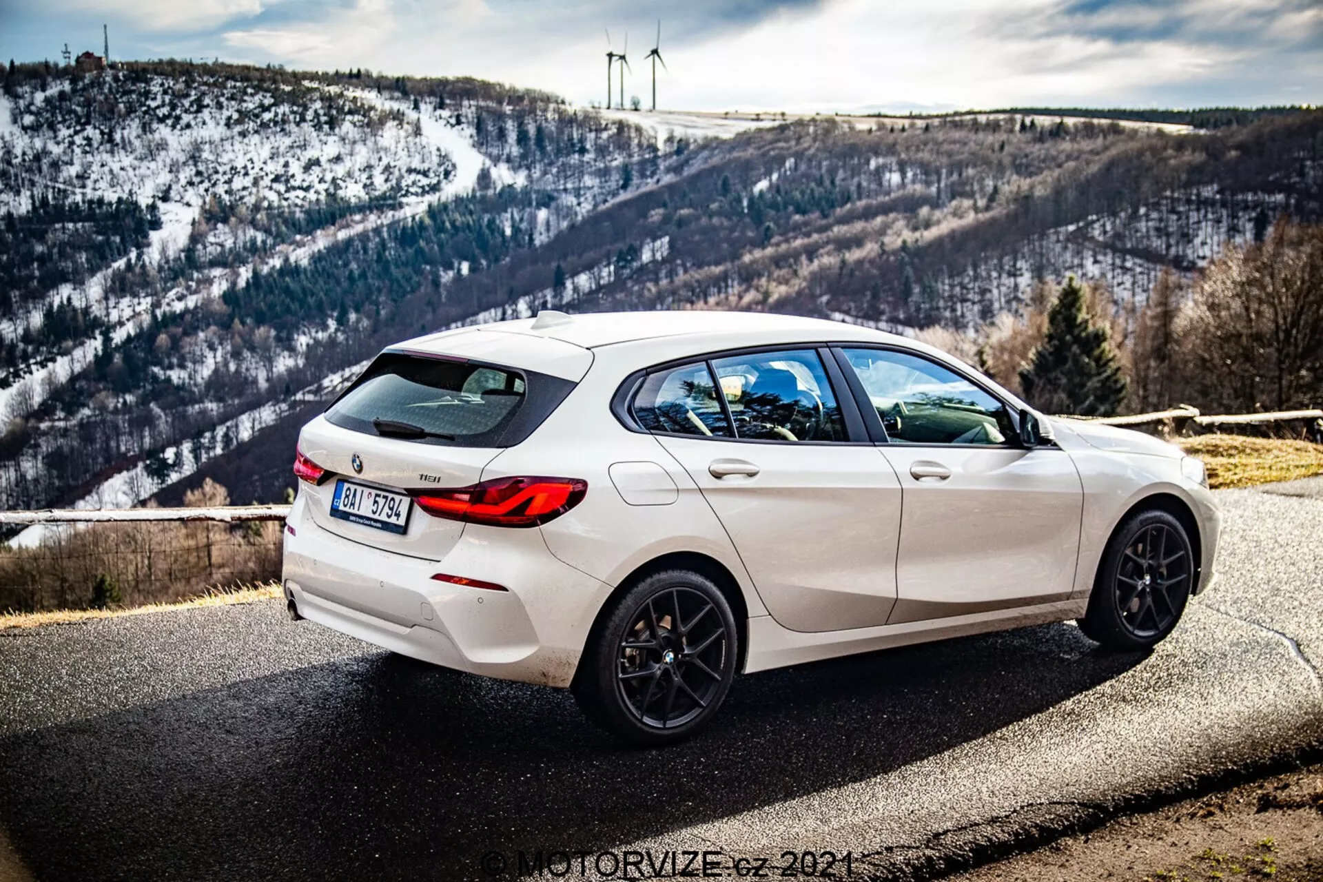 La imagen muestra un BMW Serie 1 (BMW-116i) de 2019 blanco estacionado al costado de la carretera con un fondo pintoresco que incluye un paisaje montañoso cubierto de nieve y turbinas eólicas en el horizonte. La fotografía está tomada desde un ángulo trasero de tres cuartos, mostrando prominentemente el lado derecho del coche. La vista muestra las líneas de diseño elegantes, el alerón trasero, las llantas negras de cinco radios, las salidas de escape dobles y las lunas traseras oscuras. El coche también cuenta con un limpiaparabrisas trasero y una antena tipo aleta de tiburón integrada en el techo. La profundidad de campo mantiene el coche enfocado con luz natural acentuando su estética deportiva mientras el fondo permanece sutilmente detallado.