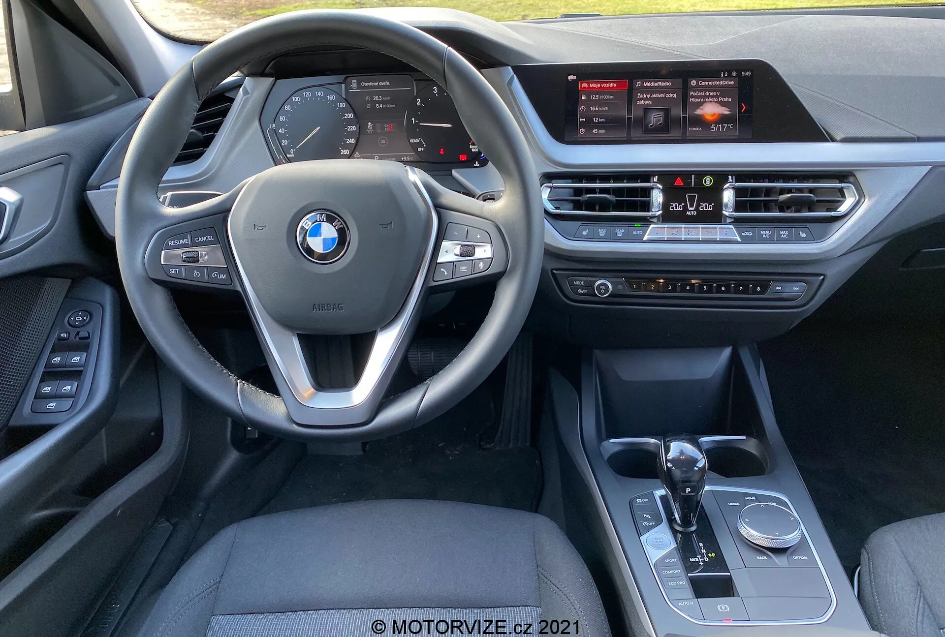 Vista interior del BMW Serie 1 (BMW 116i) 2019 desde la perspectiva del asiento del pasajero delantero, que muestra el volante multifunción con controles incorporados, el cuadro de instrumentos digital, la gran pantalla táctil de infoentretenimiento, el sistema de control de clima, la palanca de cambios automática, el selector de modo de conducción y el botón de control iDrive.