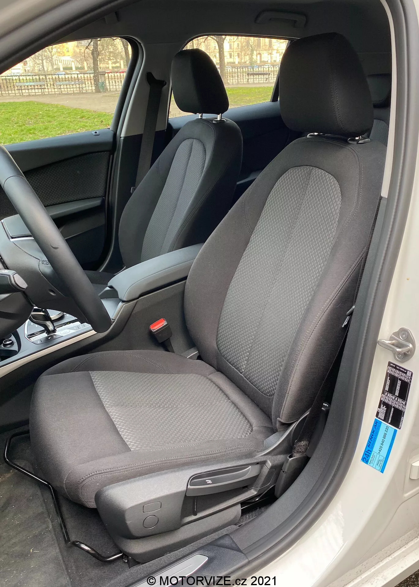 Toma de primer plano del interior del BMW 1er-Reihe 2019 (BMW-116i) centrada en el asiento del conductor con un diseño moderno de tela de dos tonos, predominantemente negro con patrones en gris claro, tomada desde un ángulo lateral con la puerta del coche completamente abierta. El asiento cuenta con un reposacabezas contra la luz, creando un efecto de silueta alrededor de los bordes, e incluye una palanca para ajustes manuales hacia adelante o hacia atrás, así como un pequeño botón negro cerca del borde del interior del coche. La luz natural del día realza el enfoque nítido en el asiento, con un fondo desenfocado que muestra el volante y el tablero de instrumentos.