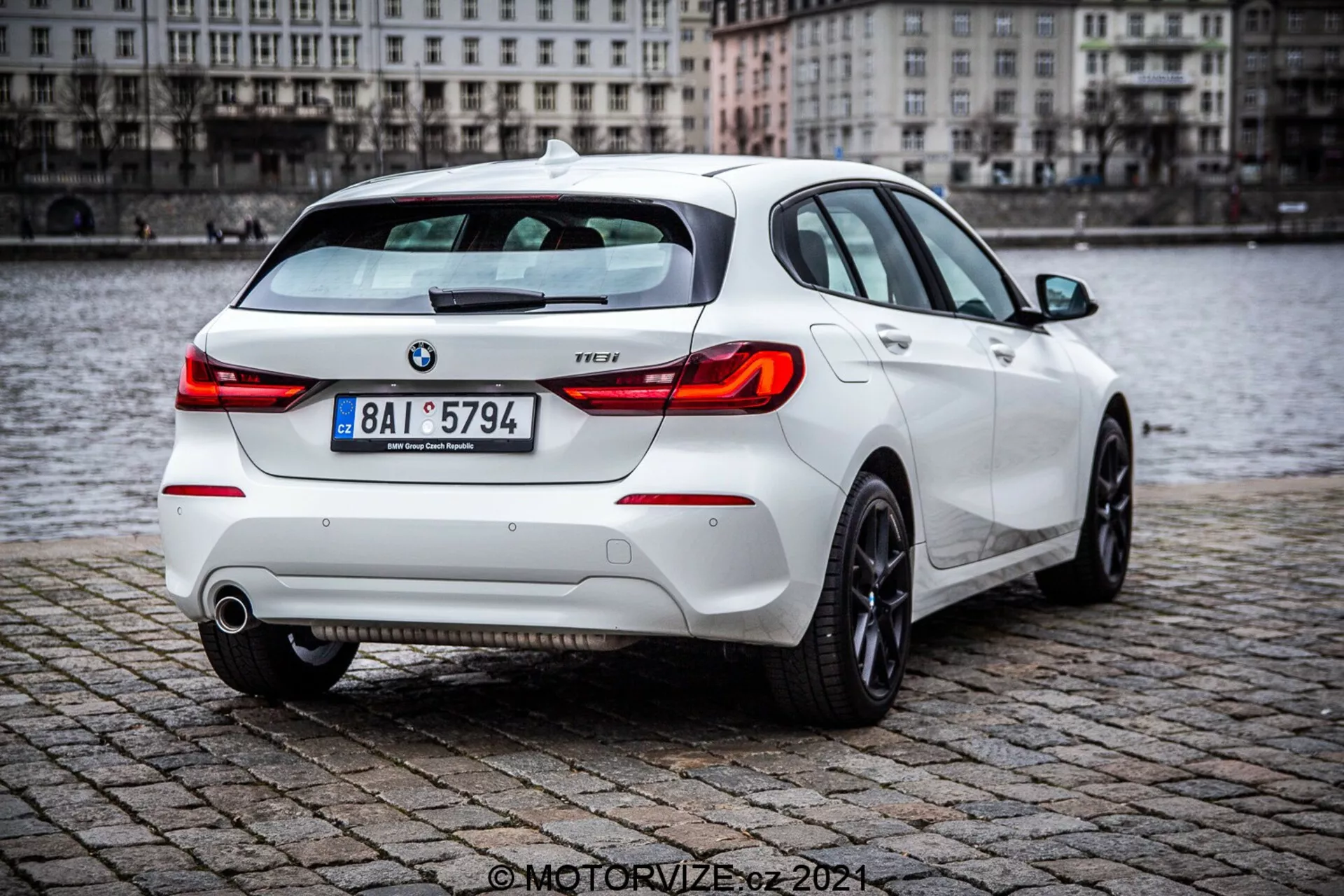 BMW Serie 1 de 2019 (116i) hatchback blanco, que muestra vista en ángulo trasero de tres cuartos con elementos detallados como luces traseras, tapa del maletero, parachoques trasero, tubos de escape y parte de las ruedas traseras, estacionado sobre superficie de adoquines mojados cerca de un cuerpo de agua. La imagen destaca el alerón trasero deportivo, los modernos conjuntos de luces traseras y las insignias visibles de BMW y nivel de equipamiento.