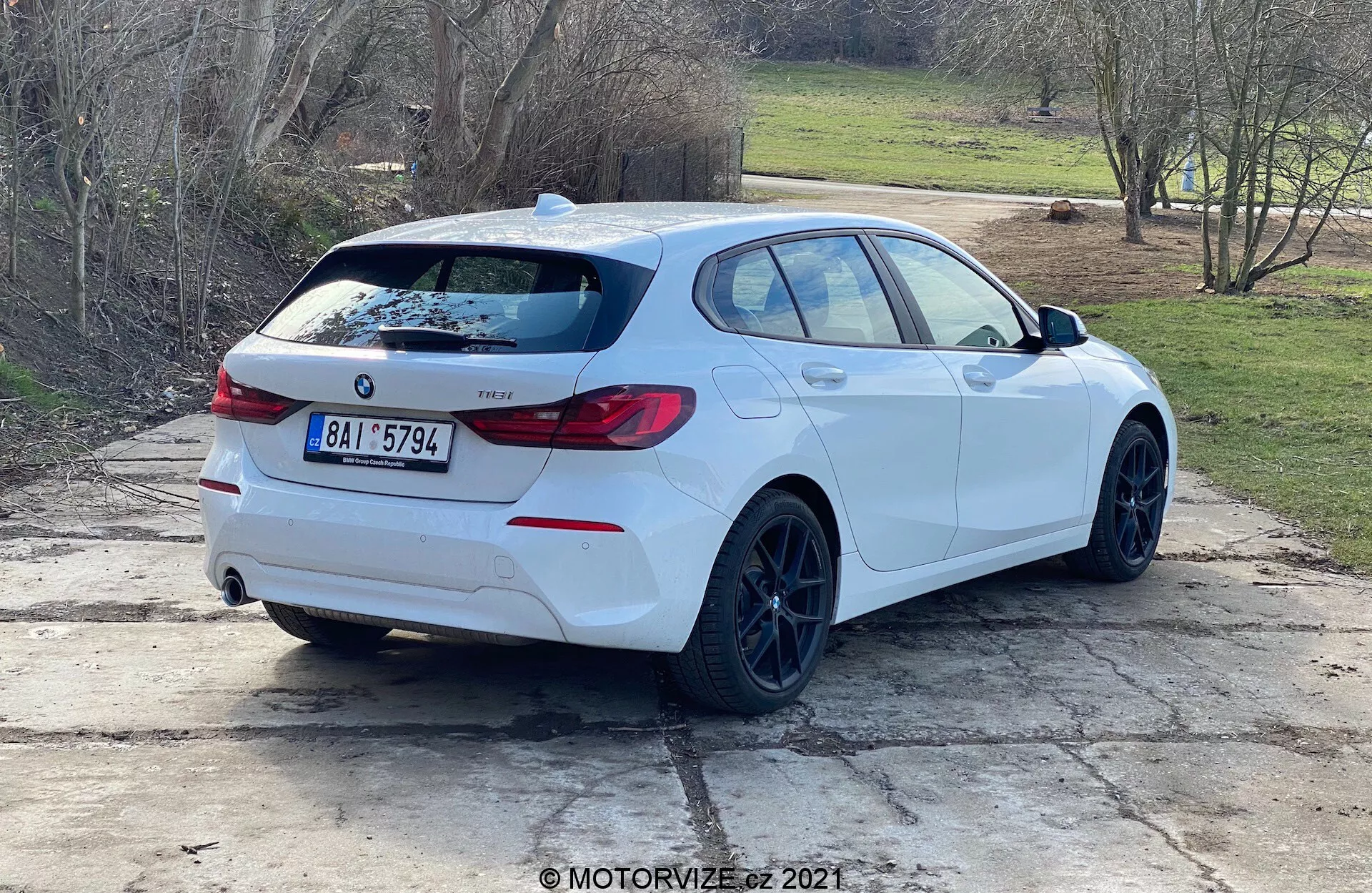 Vista trasera en tres cuartos de un BMW Serie 1 hatchback blanco (modelo BMW 116i 2019) estacionado al aire libre durante el día. Son claramente visibles el perfil lateral y la parte trasera del coche, con el emblema de BMW, la designación del modelo, las luces traseras envolventes distintivas, un alerón trasero, llantas negras, estilo de carrocería hatchback, tubos de escape dobles, limpiaparabrisas trasero, ventanas ligeramente tintadas y espejos retrovisores con indicadores de giro integrados. El coche está posicionado en un espacio de concreto con parches de hierba y un entorno rural o suburbano en el fondo.