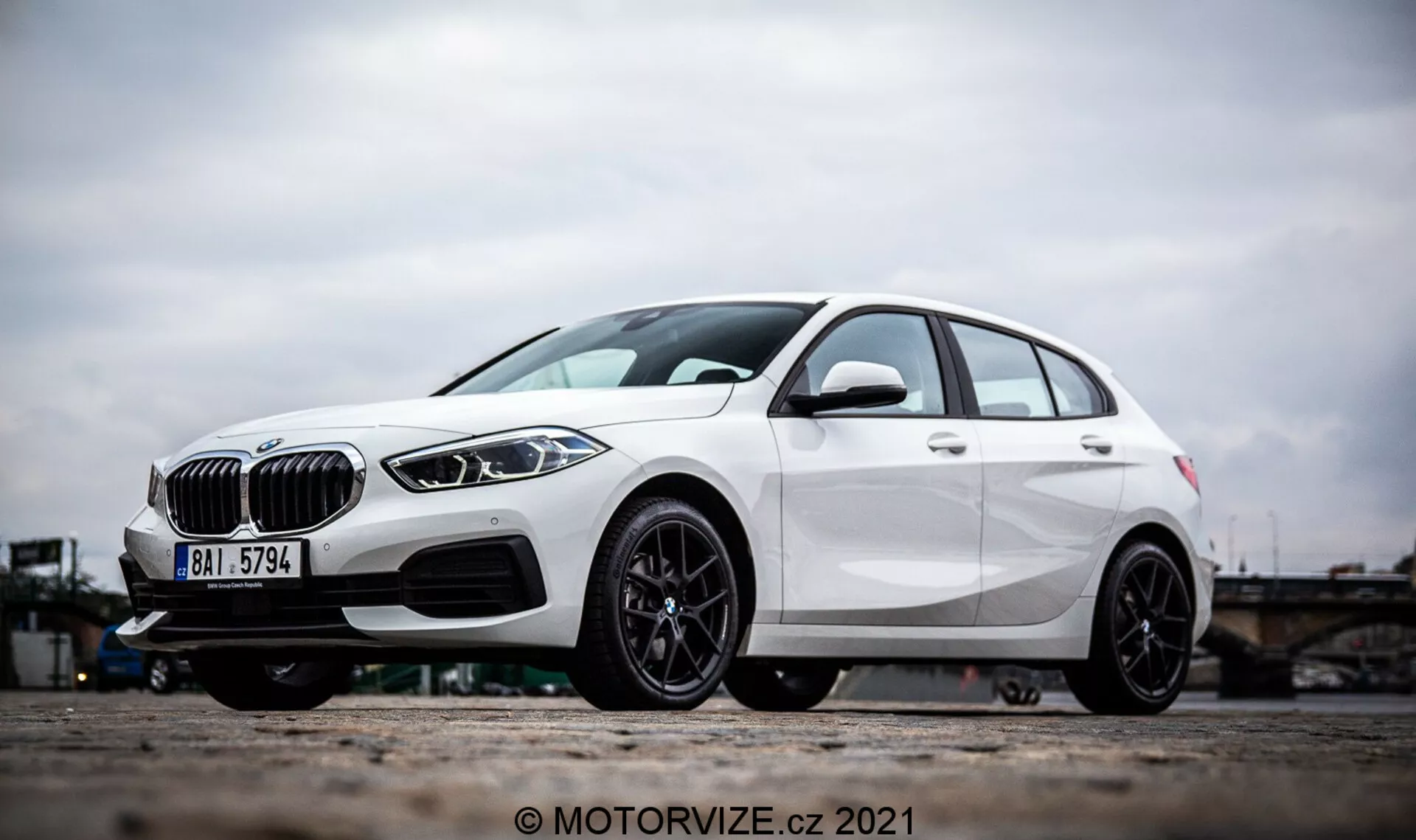 Vista lateral en ángulo de un BMW Serie 1 2019 (BMW 116i) blanco, que presenta elementos de diseño destacados como una rejilla en forma de riñón, faros LED elegantes y un parachoques delantero con tomas de aire y posibles sensores de radar para asistencia al conductor. La foto se centra en las llantas de aleación y la parte inferior del vehículo, destacando el diseño deportivo de aleación negra de múltiples radios y el freno de disco de alto rendimiento. Las líneas de carácter y la escultura aerodinámica del hatchback enfatizan aún más su exterior moderno y dinámico.