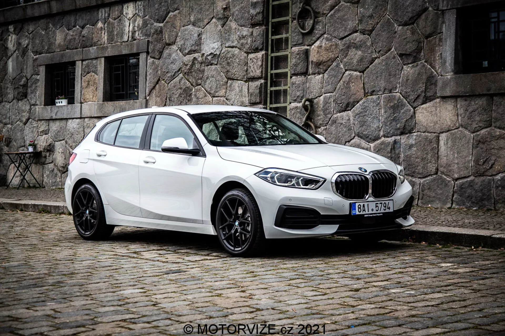 Un BMW 1er-Reihe 2019 (BMW 116i) hatchback blanco visto desde un ángulo frontal de tres cuartos, que muestra el lado izquierdo del vehículo. La foto, tomada desde un ángulo ligeramente más bajo, acentúa la postura y las líneas de diseño del coche. Está posicionado en una calle de adoquines con un edificio de piedra antigua en el fondo, creando una escena urbana clásica. La parrilla de riñón y el emblema redondo de BMW son prominentes en la parte delantera, flanqueados por elegantes faros LED angulados. El coche cuenta con llantas de aleación negras de múltiples radios, una línea de techo inclinada, espejos laterales del color de la carrocería con señales de giro integradas y ventanas tintadas para los pasajeros traseros.