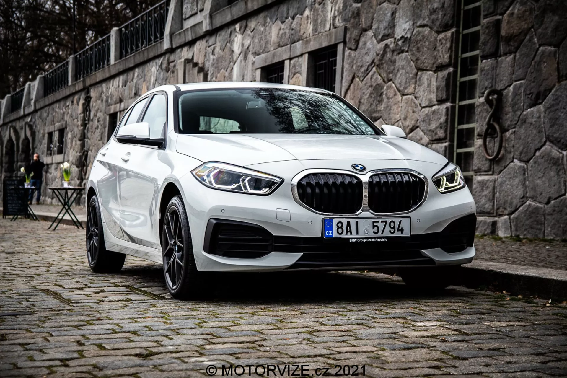 Moderno BMW Serie 1 blanco (BMW 116i) modelo 2019 que muestra la distintiva parrilla en forma de riñón, luces diurnas LED circulares dobles y diseño de parachoques delantero deportivo, estacionado sobre una superficie de adoquines con un entorno europeo histórico que presenta una pared de piedra y muebles de café al aire libre en el fondo.