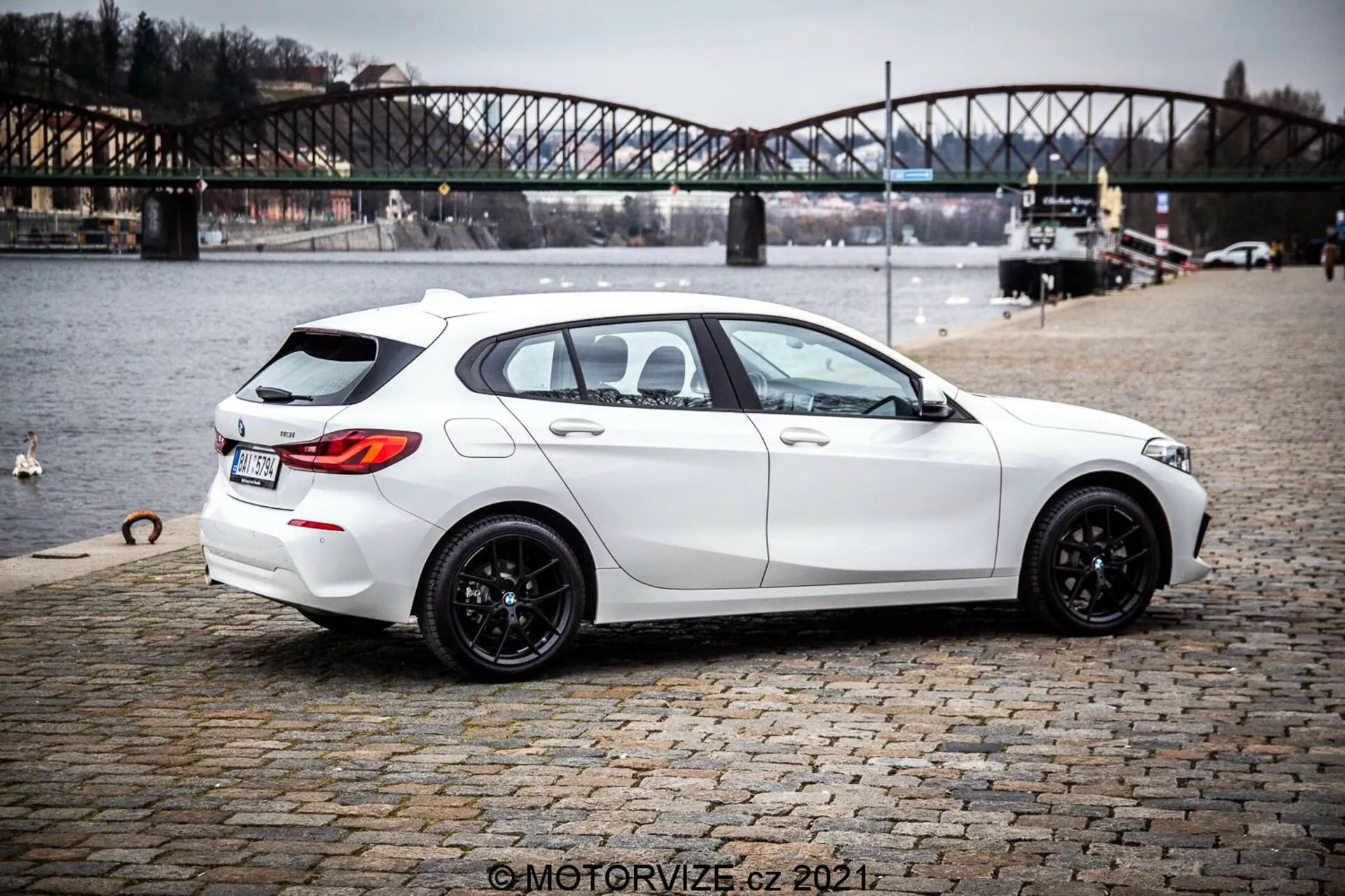 Imagen del hatchback BMW 1er-Reihe 116i blanco de 2019 desde una vista trasera de tres cuartos, destacando su diseño moderno y elegante con una postura deportiva, llantas de aleación negras de múltiples radios, ventanas ligeramente tintadas y características prácticas como el descongelador y limpiaparabrisas de la ventana trasera. El coche muestra líneas y contornos nítidos, sistema de escape dual, discreto spoiler trasero en el techo, cámara de visión trasera y sensores de aparcamiento. Está aparcado en una superficie de adoquines cerca de un cuerpo de agua con un puente urbano en el fondo, sugiriendo un contexto de estilo de vida al aire libre.
