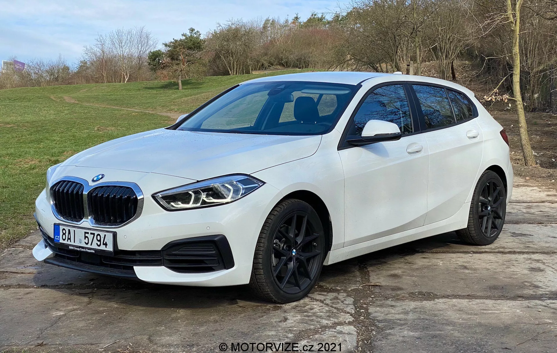 La imagen muestra un moderno hatchback BMW Serie 1 blanco (BMW 116i) ubicado al aire libre con un fondo natural y de hierba. Capturado desde una vista tres cuartos delantera favoreciendo el lado del conductor, destaca la parrilla distintiva del vehículo, faros LED angulares elegantes, ruedas de aleación negras, pliegue lateral bien definido y señales de giro integradas en los espejos laterales. El parachoques delantero presenta sensores de estacionamiento, mientras que se puede inferir un techo de cristal panorámico y una posible cámara frontal. El coche está estacionado en un área de adoquines y ocupa la mayor parte del cuadro, sugiriendo un diseño automotriz moderno y características tecnológicas avanzadas.