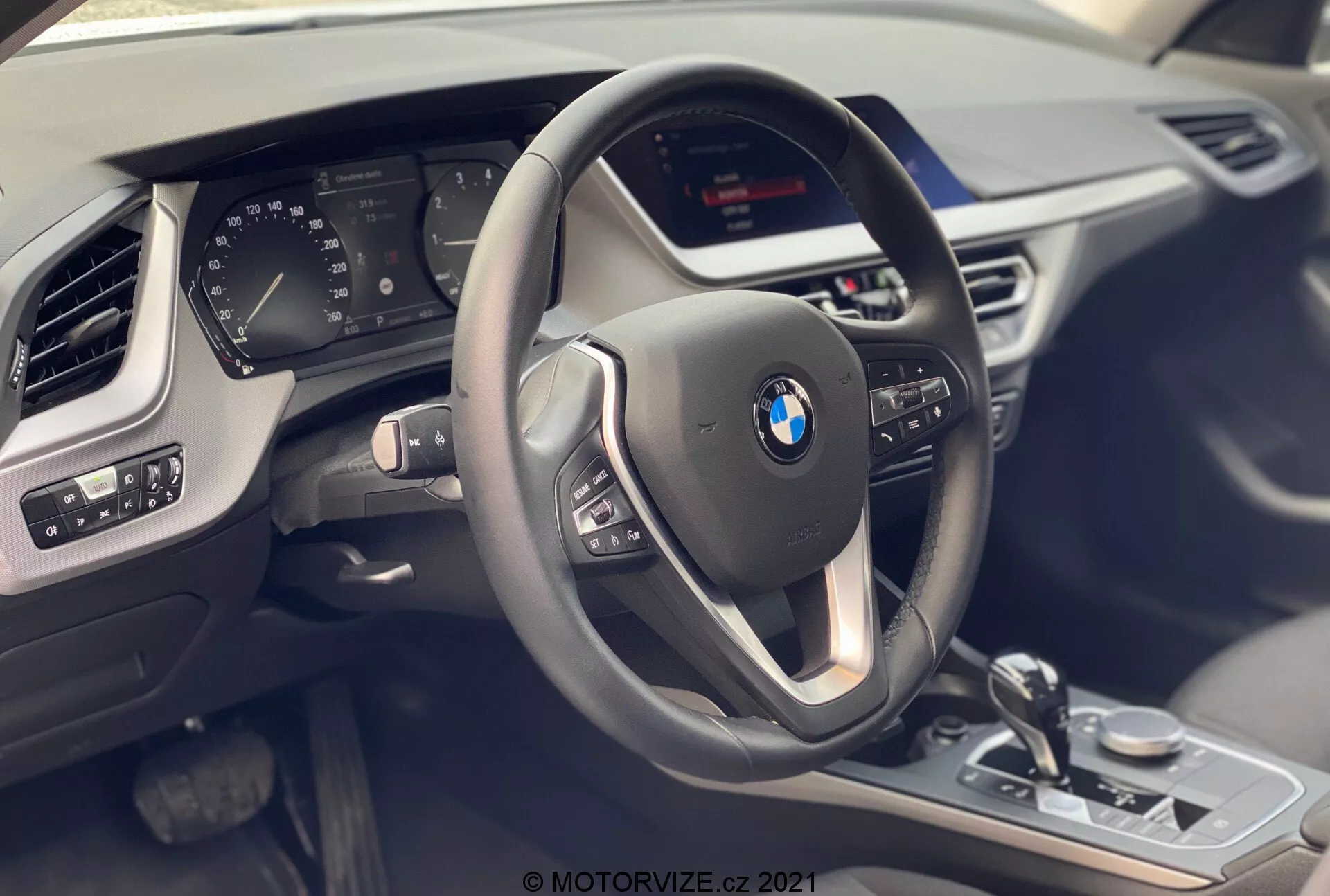 Vista interior del BMW Serie 1 2019 (BMW 116i) desde el asiento del pasajero, destacando el cockpit del conductor con un volante moderno de tres radios con botones multifunción y cubierta de cuero con detalles de costura contrastante. Detrás del volante hay un cuadro de instrumentos digital parcialmente visible que muestra varios parámetros del vehículo. La consola central alberga una pantalla de infoentretenimiento no visible y controles visibles como la palanca selectora de cambios, el botón de control iDrive, el selector de modo de conducción y los controles del sensor de estacionamiento, todos con acabados metálicos y moldura negra brillante. El panel de la puerta del conductor muestra ajustes de espejos y control de ventanas eléctricas. También se ve una parte del ventilador del lado del pasajero y la textura del tablero, subrayando la sensación de lujo, tecnología y diseño moderno.
