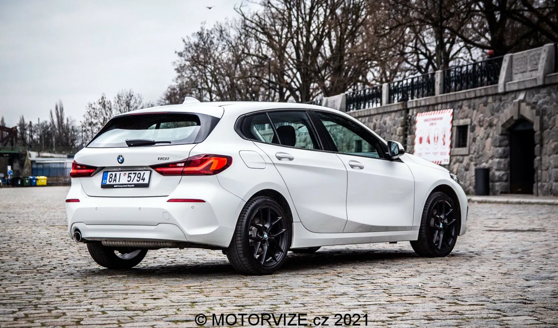 Una fotografía de alta calidad de un hatchback BMW de la Serie 1 2019 128ti blanco tomada desde un ángulo trasero en tres cuartos, destacando el lado derecho. El coche está aparcado en un pavimento adoquinado contra un fondo de pared de piedra, enfatizando su diseño deportivo con un alerón trasero, sistema de escape doble, difusor trasero, luces traseras LED, ventanas tintadas y ruedas de aleación de color oscuro. La iluminación difusa, la ausencia de sombras duras y los detalles claros sugieren un ambiente nublado, proporcionando una representación auténtica del color y las características del vehículo. La imagen tiene una marca de agua 'MotorVize.cz 2021'.