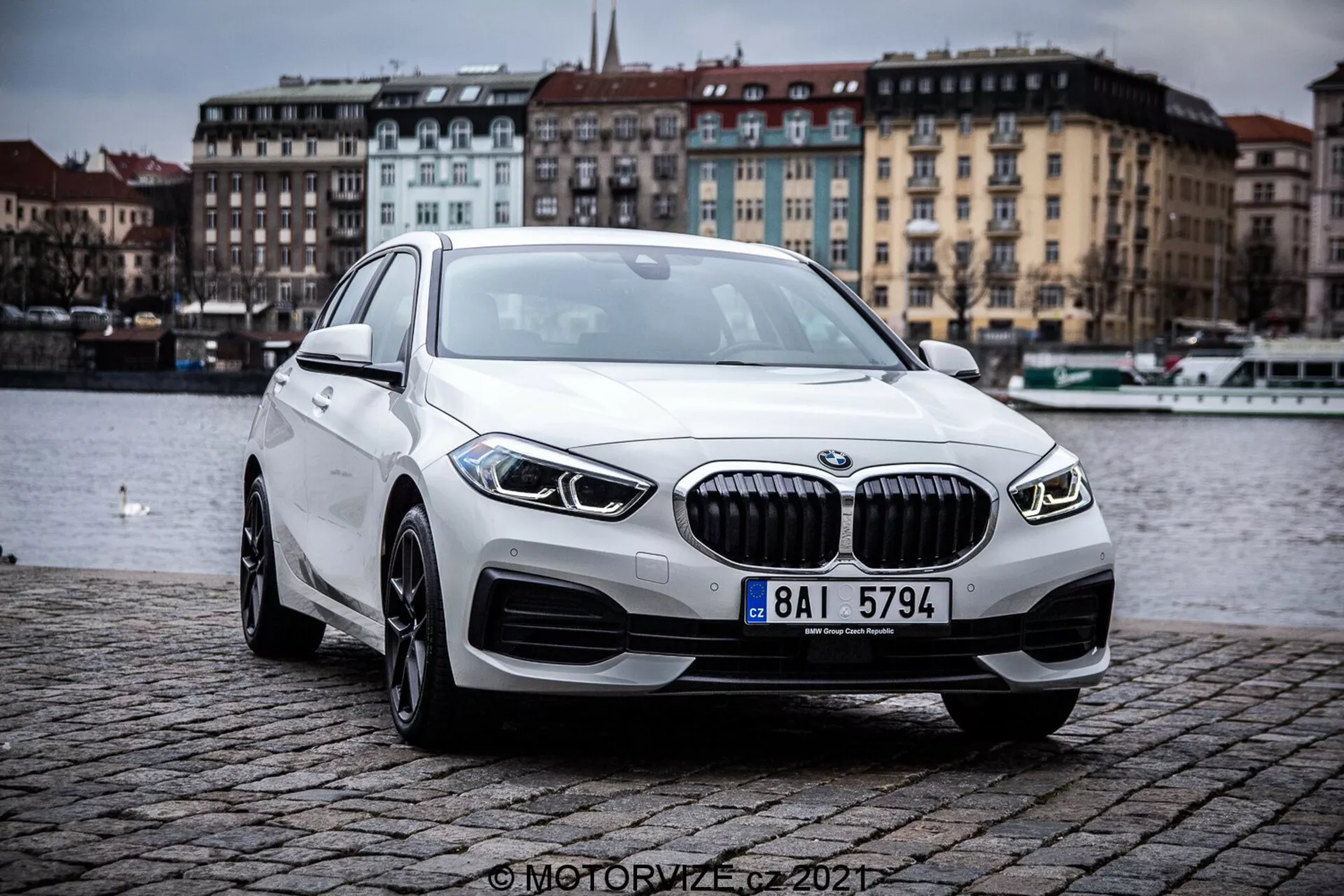 Imagen que muestra un BMW 1er-Reihe 2019 (BMW 116i) desde una perspectiva frontal tres cuartos, resaltando los contornos y elementos de diseño del sedán de lujo moderno. El coche se encuentra al aire libre en un terraplén empedrado cerca de un cuerpo de agua, bajo un cielo nublado. Las características prominentes incluyen una parrilla con láminas verticales, elegantes faros LED, luces antiniebla integradas, ruedas de aleación y perfil aerodinámico.