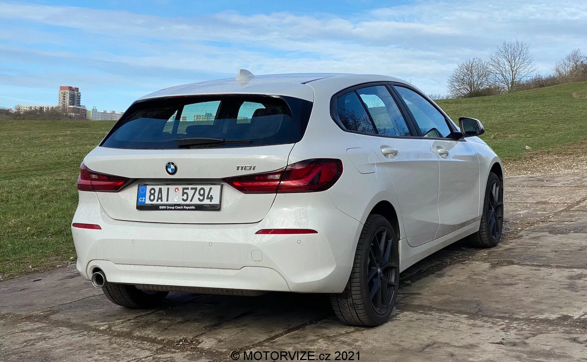 Vista trasera de tres cuartos de un hatchback BMW 1 Series 118i blanco, estacionado en un terreno irregular. El coche cuenta con luces traseras distintivas en forma de 'L', un emblema de BMW y una insignia de modelo que dice '118i'. Tiene un sistema de escape doble con una tubería visible en el lado derecho del parachoques trasero. El vehículo está equipado con ruedas de aleación negras de múltiples radios y un limpiaparabrisas trasero. Un alerón en el techo añade al aspecto deportivo, y el perfil lateral revela ventanas ligeramente tintadas, pliegues en la carrocería y manijas y espejos que coinciden con el esquema de color del coche. El ángulo bajo de la cámara enfatiza la postura del coche y el diseño de las ruedas.