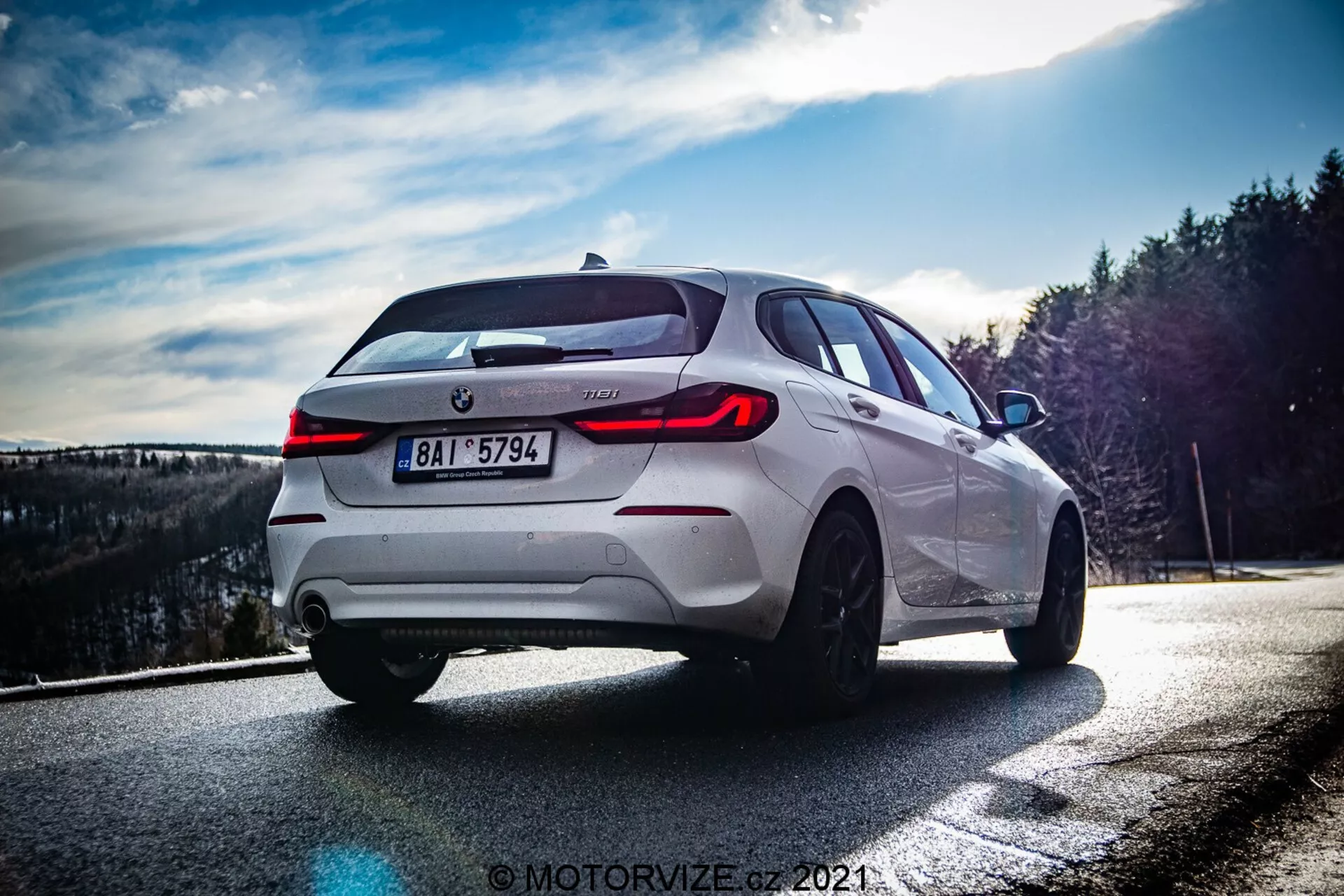 La imagen muestra un BMW Serie 1 2019 blanco (BMW-116i) capturado desde un ángulo bajo y algo trasero de tres cuartos, enfatizando el lado derecho trasero del coche. La configuración diurna, probablemente al final de la tarde, crea contrastes dinámicos que resaltan las líneas de diseño. Las características prominentes incluyen luces traseras LED distintivas, emblema del fabricante y badging del modelo centrados, sistema de escape dual, llantas de aleación de múltiples radios, neumáticos de bajo perfil, frenos de disco visibles y una silueta deportiva de hatchback con parabrisas trasero inclinado y alerón de techo. La composición y los efectos de iluminación de la fotografía añaden un toque dramático, con el fondo borroso para enfocarse únicamente en el vehículo.