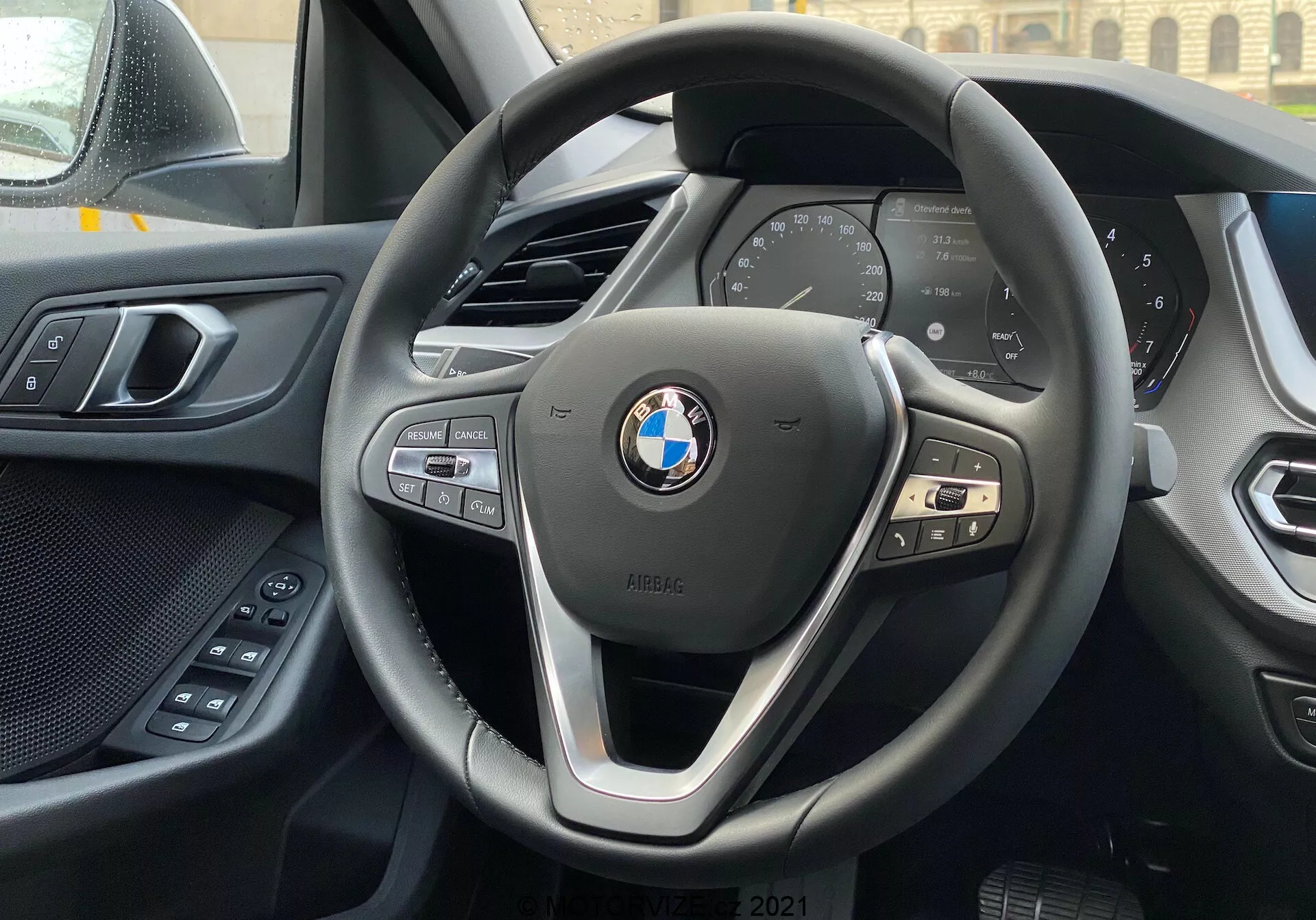 Vista interior de un BMW Serie 1 2019 (BMW 116i) desde el asiento del conductor, centrada en el volante moderno de tres radios de BMW con botones de control para multimedia, teléfono y control de crucero, y el cuadro de instrumentos digital que muestra métricas esenciales del vehículo como velocidad y RPM. El borde superior de la consola central es visible en el fondo, y a la izquierda se ve parte de la puerta del conductor con controles eléctricos de confort. El tablero y el panel de la puerta muestran una combinación de materiales suaves al tacto y una fabricación de calidad. El emblema de BMW y la insignia iPERFORMANCE se muestran prominentemente en el volante, lo que sugiere eficiencia o tecnología híbrida.
