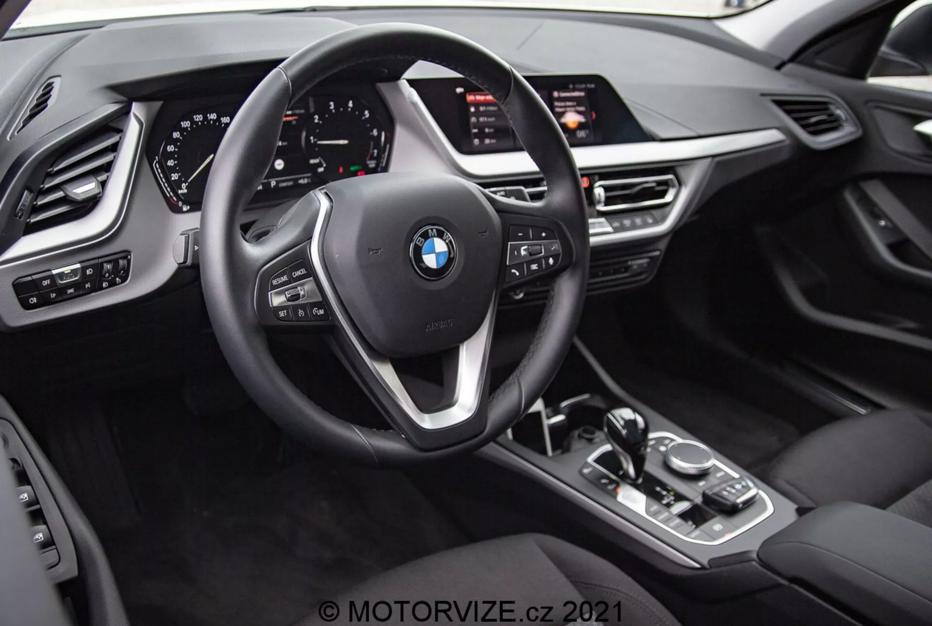 Vista interior del BMW 1er-reihe 2019 (BMW 116i) desde la perspectiva del conductor. La imagen destaca el emblema de BMW en el volante de tres radios con botones multifuncionales y paletas de cambio detrás del volante. Detrás del volante se encuentra un cuadro de instrumentos completamente digital con diales iluminados y varias pantallas informativas. La consola central cuenta con una palanca de cambios automática, un mando de control iDrive, botones para los modos de conducción y una gran pantalla de infoentretenimiento inclinada hacia el conductor. El diseño interior incluye texturas similares al cuero, acentos metálicos y controles adicionales para luces, ventanas y ajustes de espejos con configuración de memoria para el asiento del conductor.