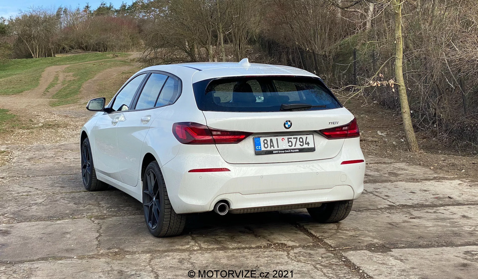 La imagen muestra un BMW 1er-reihe 2019 (BMW 116i) blanco hatchback visto desde un ángulo trasero de tres cuartos. La parte trasera del vehículo, incluyendo las luces traseras orientadas horizontalmente, el sistema de doble escape y los sensores de aparcamiento integrados en el paragolpes trasero, son claramente visibles. El emblema y el distintivo del modelo BMW están en el portón trasero. El coche está situado sobre una superficie de grava con un fondo natural y rural. La imagen sugiere temas automovilísticos como diseño de vehículos, modelos contemporáneos, contenido específico de BMW, sistemas de escape y tecnología de asistencia de aparcamiento. La marca de agua MOTORVIZE.cz 2021 indica un origen en una revisión o publicación automotriz.