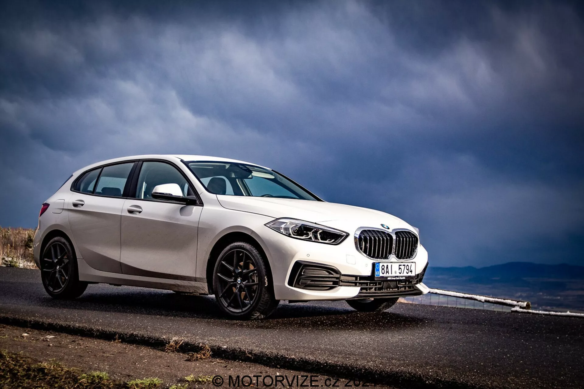 BMW Serie 1 2019 (BMW-116i) capturado en una vista frontal de tres cuartos, mostrando su perfil frontal y lateral. El coche, terminado en blanco brillante con acentos negros contrastantes como carcasas de espejos, molduras de ventanas y llantas de múltiples radios, está posicionado en el lado izquierdo del encuadre contra un fondo de una carretera asfaltada que se extiende en la distancia y un cielo tormentoso. Las características visibles incluyen faros detallados con elementos circulares, parrilla de riñón prominente, parachoques delantero con rejillas de ventilación y posibles sensores de estacionamiento.