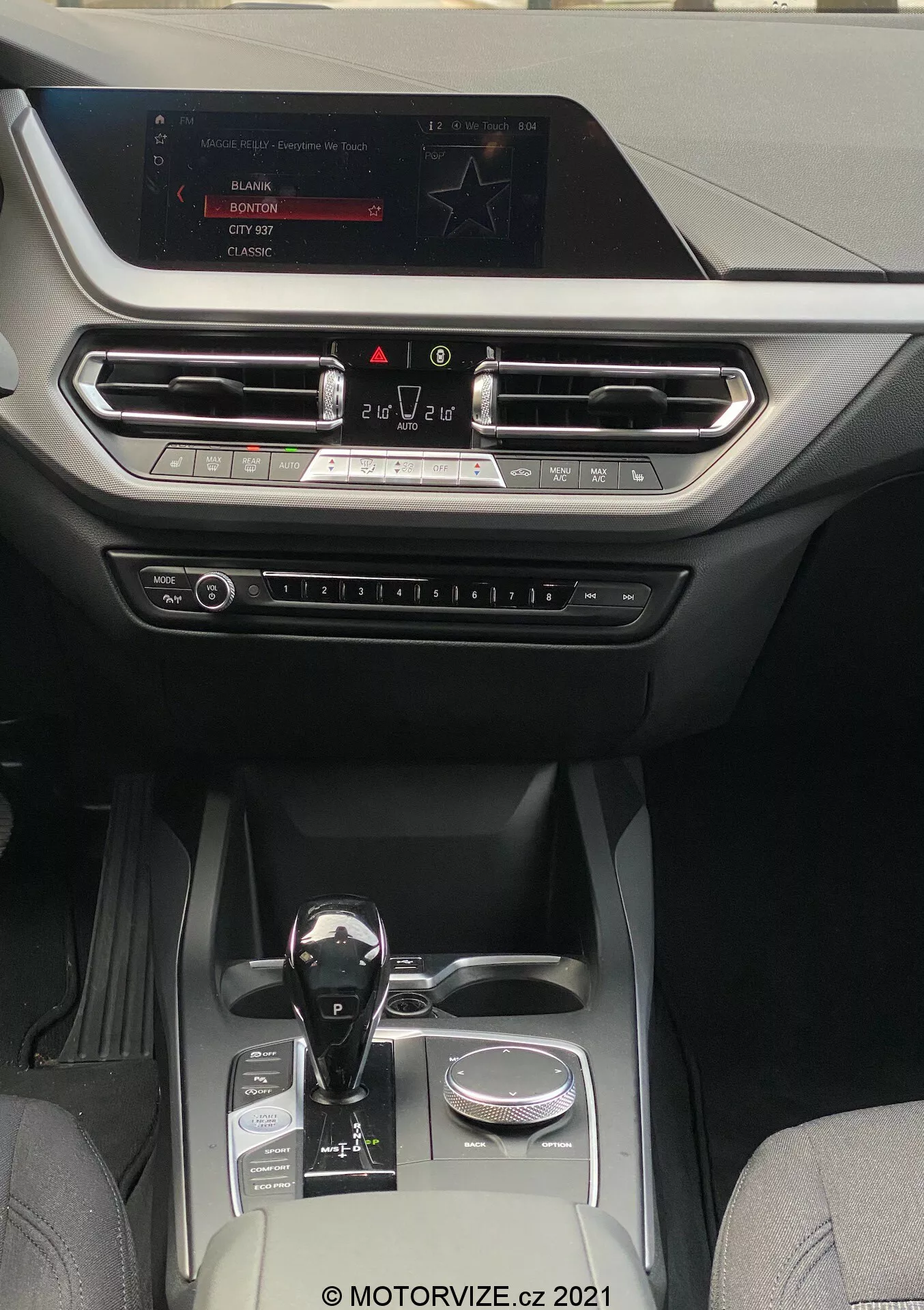 Vista interior de un BMW 1er-reihe 2019 (BMW-116i), que muestra una cabina moderna y lujosa centrada en la consola central y el área del tablero. La perspectiva es desde el área de los asientos traseros, mirando hacia adelante hacia la pantalla de infoentretenimiento montada en la parte superior del tablero. La pantalla muestra opciones de radio, medios y configuraciones del coche. Debajo de la pantalla, las rejillas de ventilación alineadas horizontalmente con acentos metálicos y una moldura decorativa se extienden a lo largo del tablero. Debajo de las rejillas de ventilación, se pueden ver botones y perillas físicas para el control del clima y otros ajustes. La palanca de cambios de transmisión automática presenta un acabado en negro brillante con acentos metálicos, posicionada al lado de un dial giratorio. La consola central incluye dos portavasos, un pequeño compartimento de almacenamiento y un elemento decorativo plateado, lo que indica una cabina bien equipada que combina cuero y telas sintéticas de alta calidad, adecuada para categorías de vehículos de rango medio a alto que se centran en la tecnología y la comodidad.