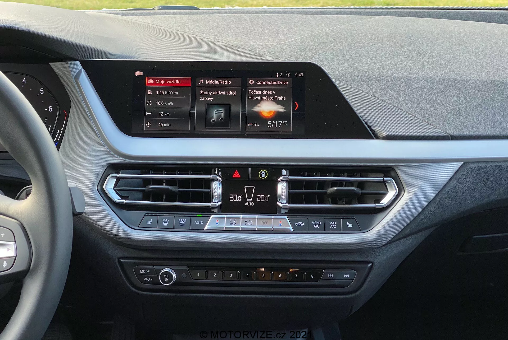 Vista interior de un BMW serie 1 de 2019 (BMW-116i) desde la perspectiva del conductor, mostrando la consola central del tablero. En el centro se encuentra la pantalla táctil rectangular e iluminada del sistema de infoentretenimiento, que muestra navegación, multimedia y detalles del clima. Debajo de la pantalla hay rejillas de ventilación con ribetes metálicos, controles de emergencia y recirculación de aire, seguidos por un panel de control del clima con botones de temperatura, velocidad de ventilador y asientos calefaccionados. Los materiales cuentan con superficies suaves al tacto y acentos cromados, sugiriendo un enfoque en la comodidad del conductor y pasajero delantero.