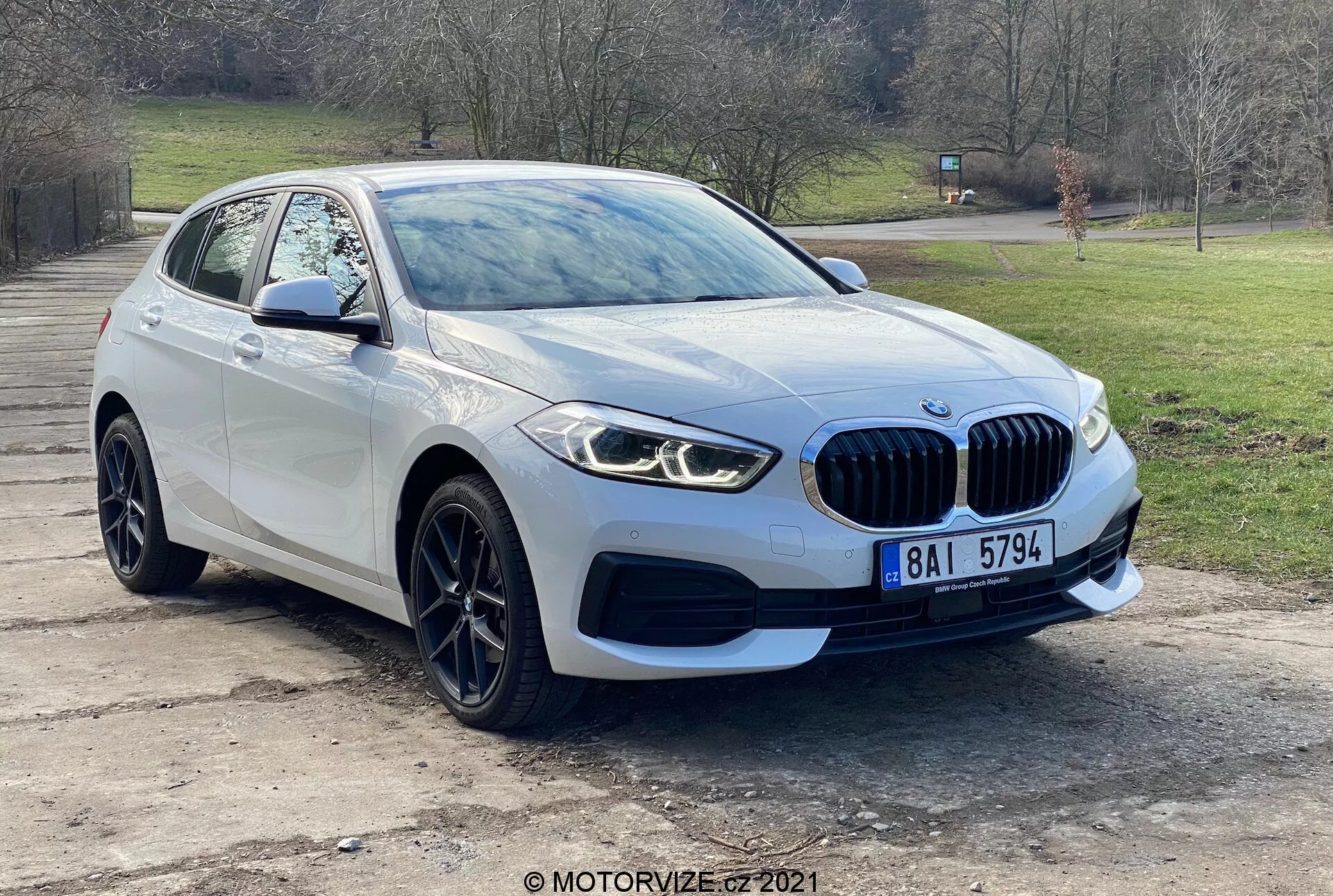 Vehículo moderno BMW 116i de la Serie 1 de 2019 fotografiado desde una perspectiva frontal de tres cuartos, mostrando la parrilla delantera con emblema, elegantes faros LED, luces antiniebla, parachoques delantero y líneas aerodinámicas del capó que conducen al parabrisas. La rueda y el guardabarros del lado del conductor, junto con el espejo lateral con intermitentes integrados, resaltan el diseño avanzado y la tecnología de sensores de aparcamiento del vehículo. Capturado al aire libre con un fondo de árboles y césped, el coche presenta llantas de aleación y una distintiva parrilla en forma de riñón, lo que indica lujo y rendimiento.